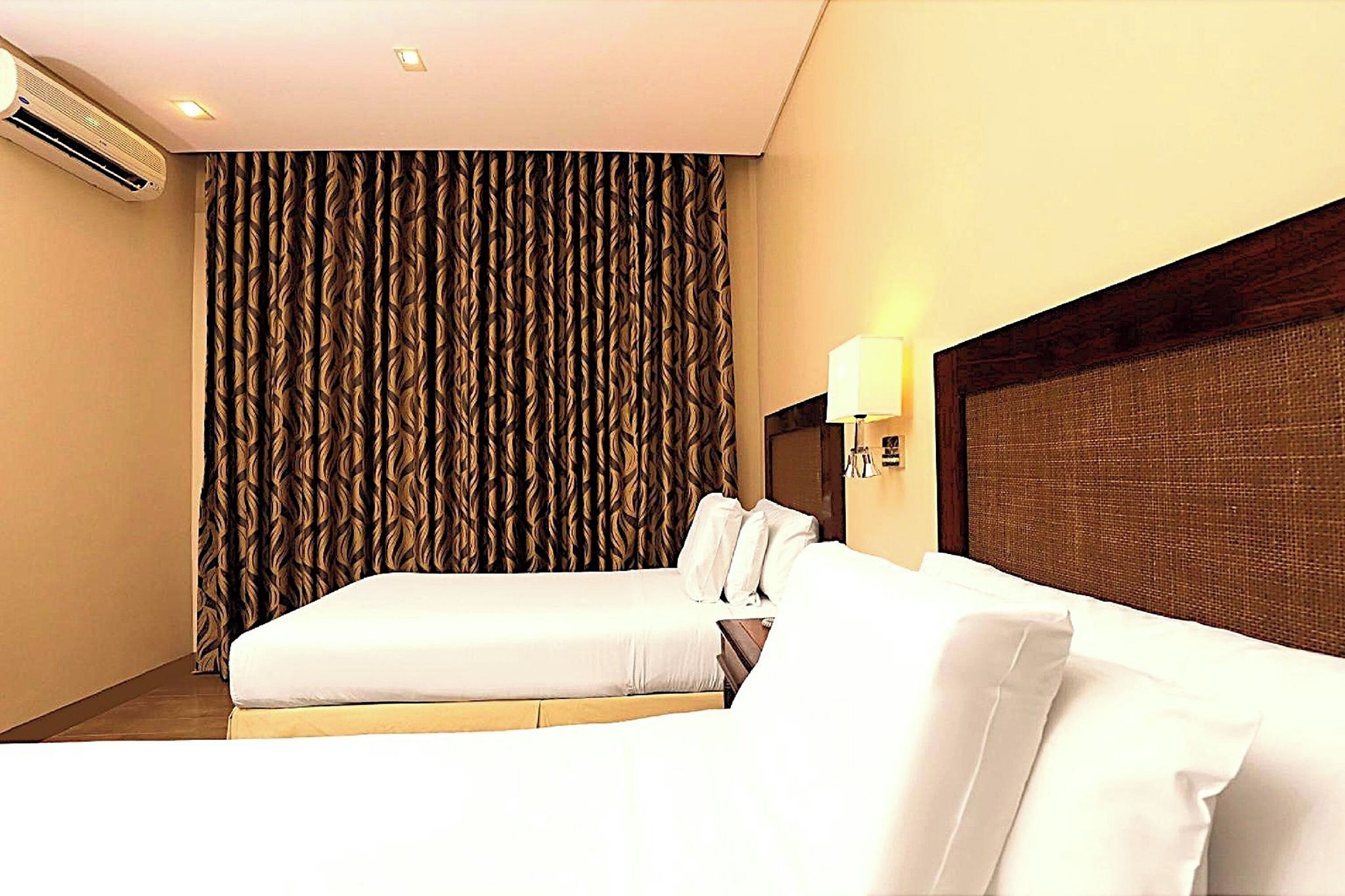 A hotel room with two beds, a large window with curtains, an air conditioning unit, and a wall-mounted lamp.