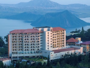 A multi-story hotel with balconies, situated near a body of water and mountains.