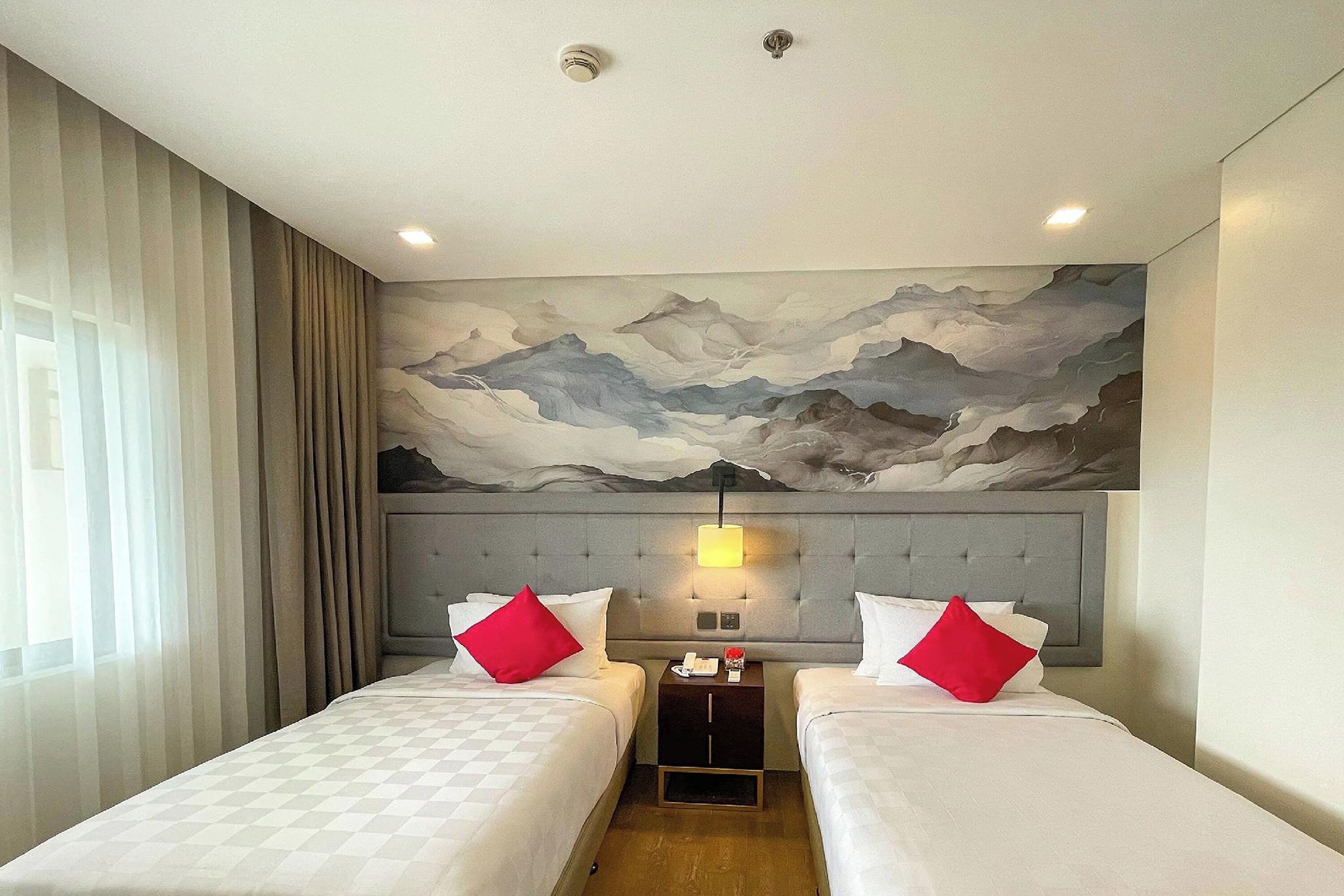 A hotel room with two beds, a headboard with a painting, a nightstand with a lamp, and a window with curtains.