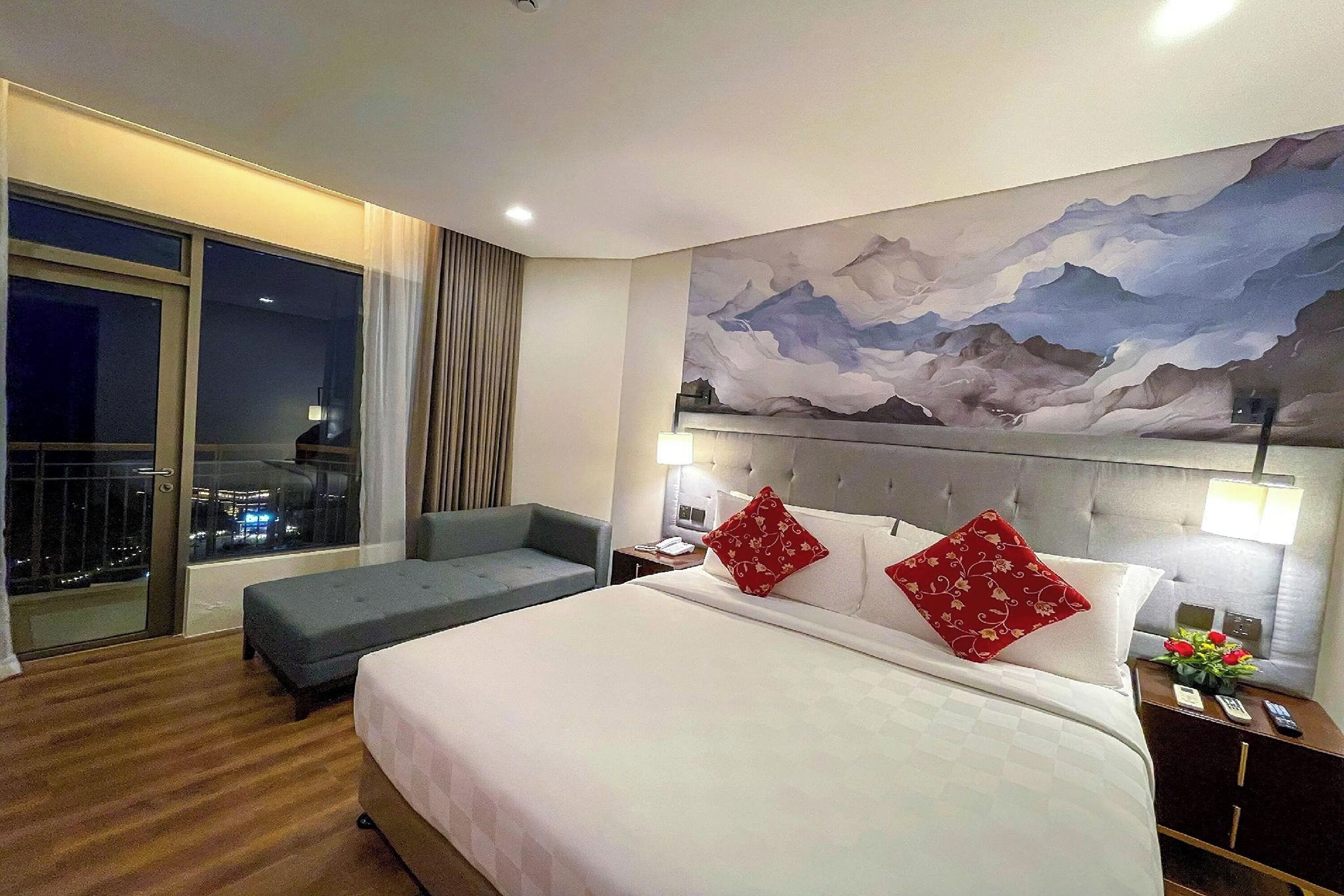 A hotel room with a large bed, a sofa, a painting of mountains, a balcony with a city view, and a night sky visible through the window.