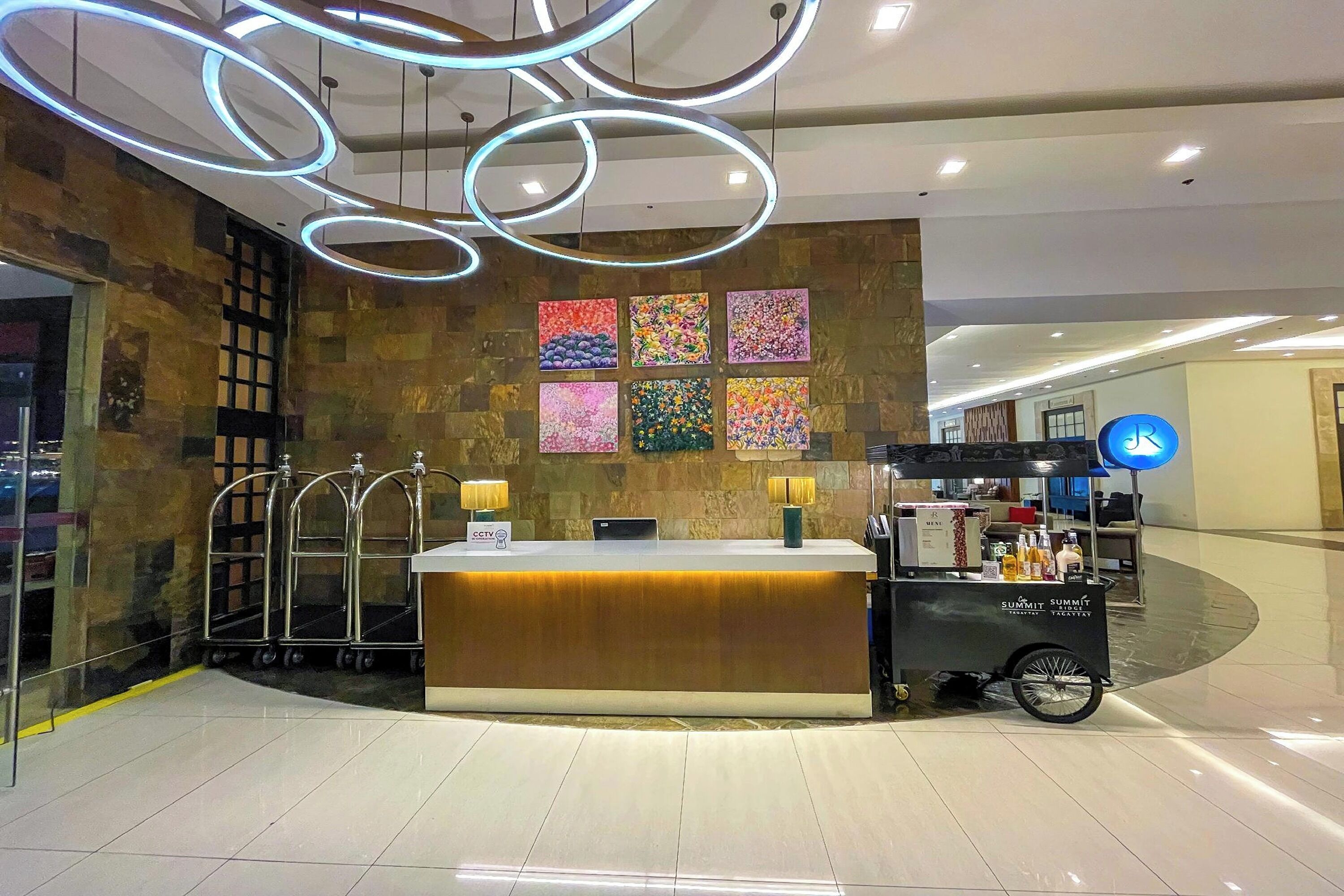 A modern hotel reception area with a central counter, a cart with beverages, and a wall with colorful artwork.