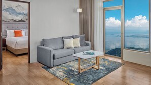 Habitación de hotel moderna con una cama grande, un sofá gris, una mesita de centro pequeña y vistas al mar a través de un ventanal amplio.