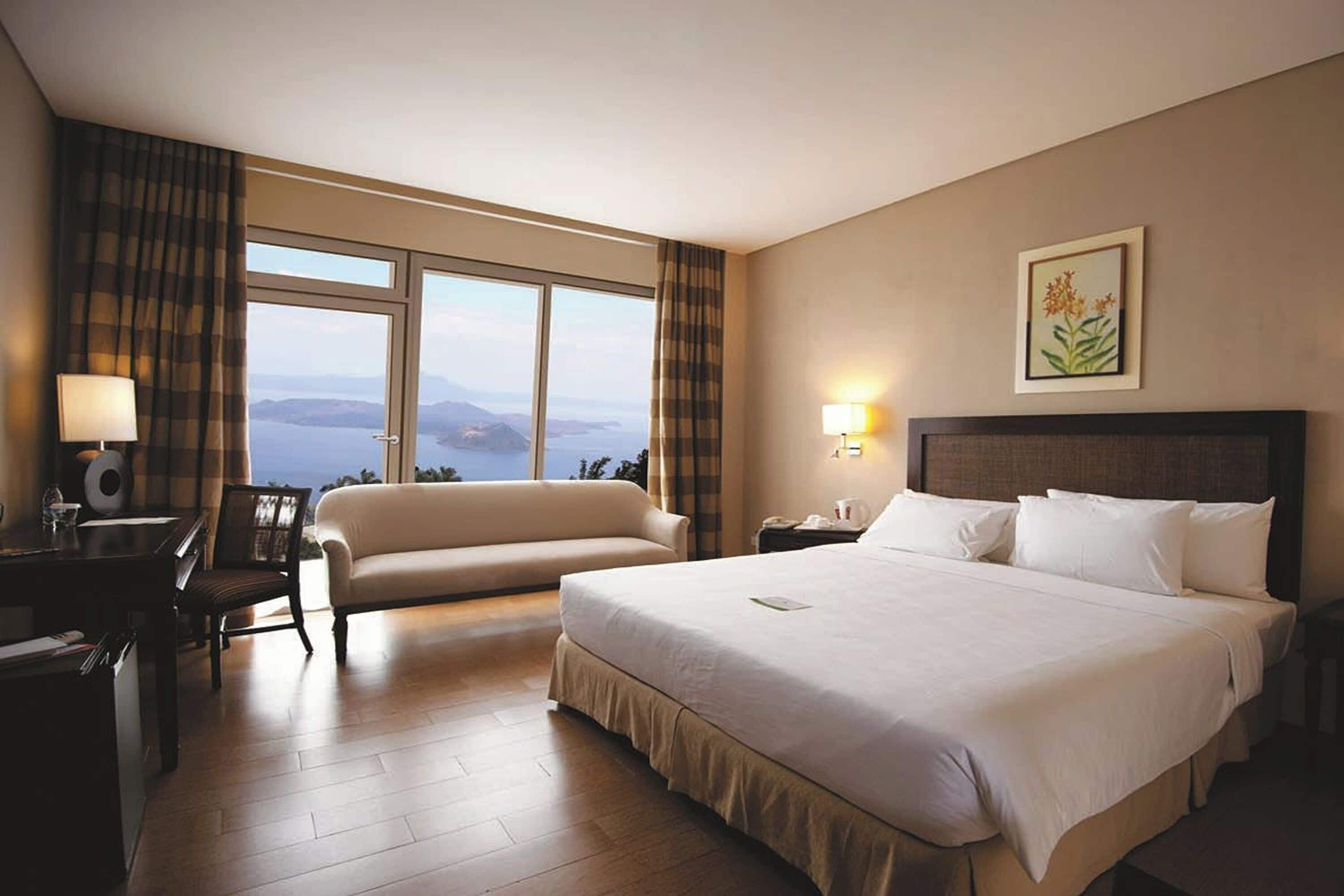 Habitación de hotel con una cama grande, un sofá, un escritorio con lámpara y vista al mar.