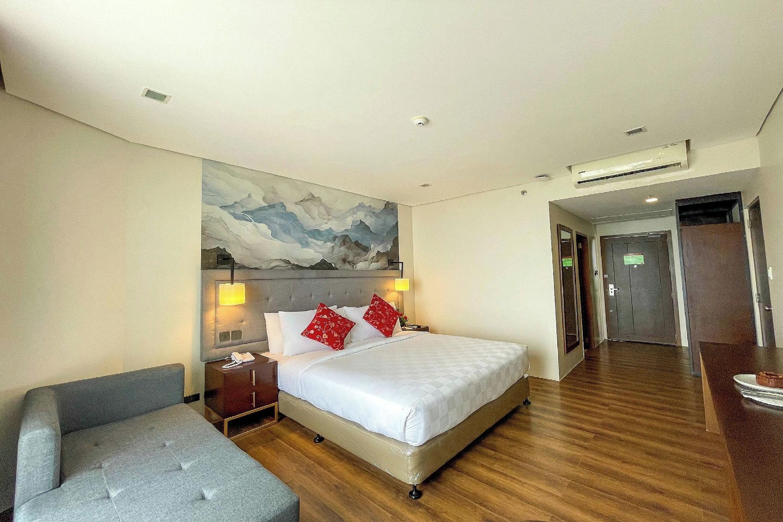 A hotel room with a large bed, a bedside table with a lamp, a grey armchair, and a painting on the wall.