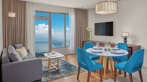 Un salón moderno con un sofá, una mesa de comedor con sillas azules, un televisor montado en la pared y un amplio ventanal con vistas panorámicas.