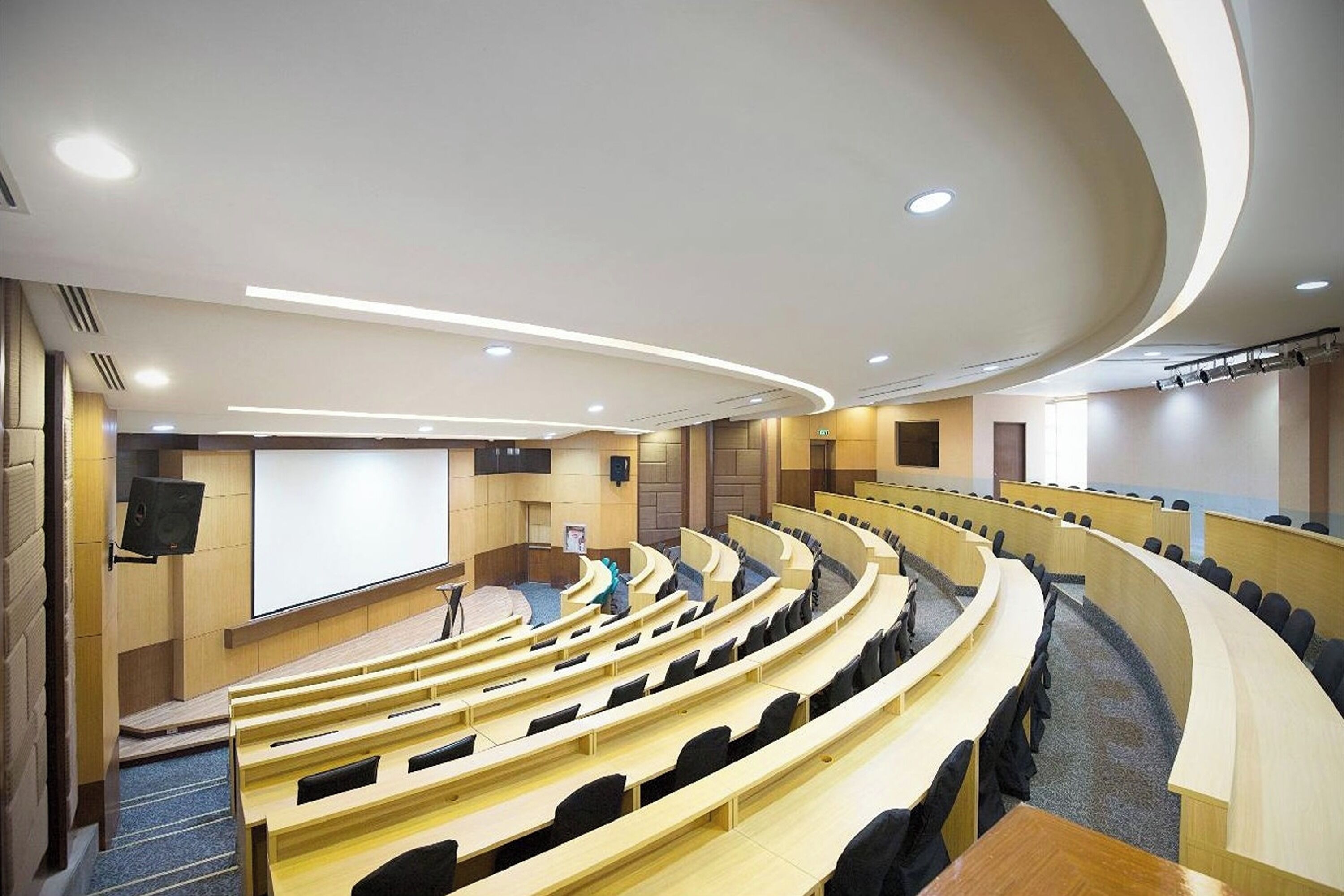 Ein modernes Auditorium mit terrassenförmigen Sitzplätzen, einer großen Leinwand und eingelassener Beleuchtung.