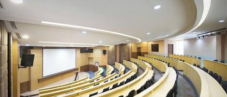 Ein modernes Auditorium mit terrassenförmigen Sitzplätzen, einer großen Leinwand und eingelassener Beleuchtung.