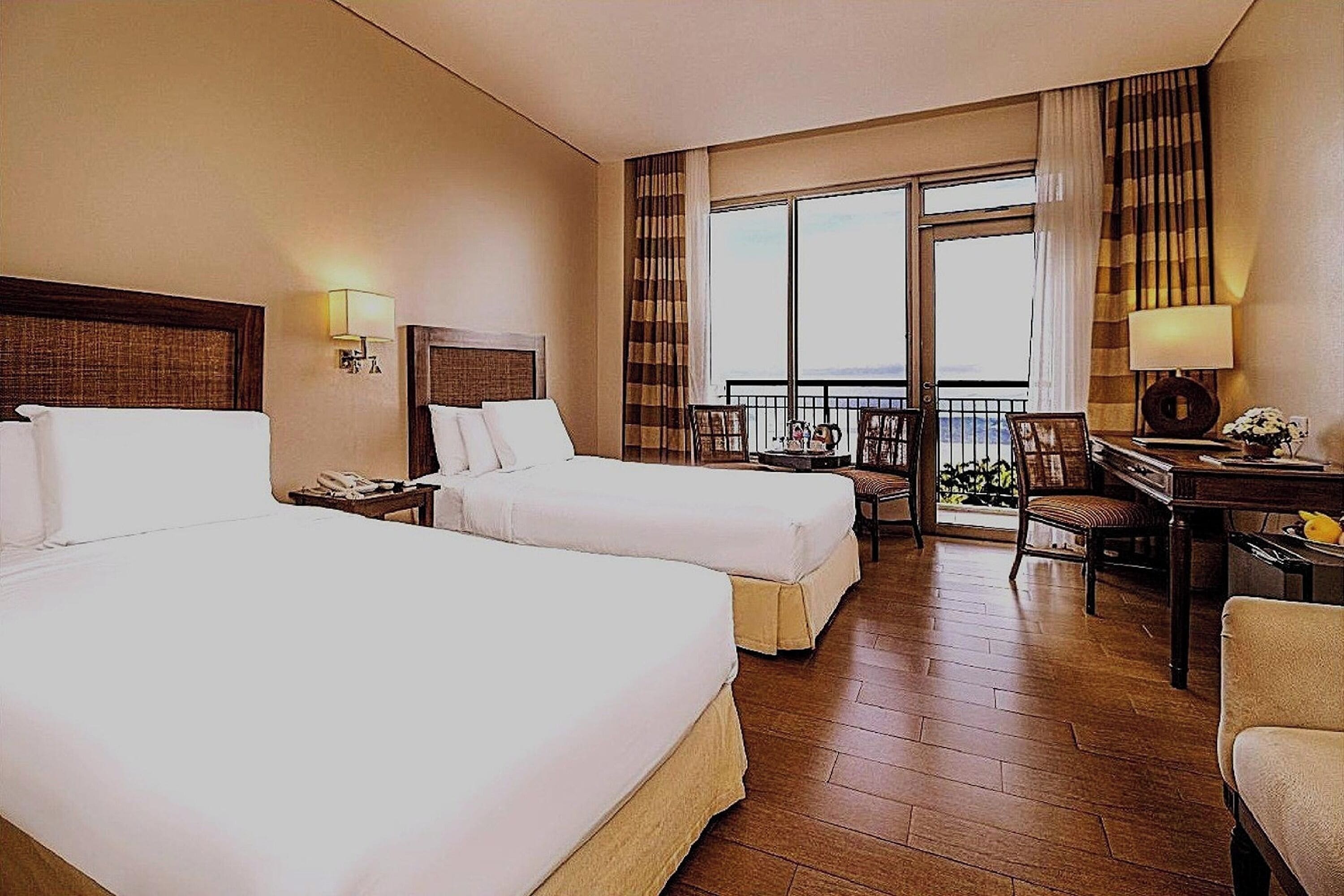 A hotel room with two beds, a desk, a chair, a balcony with a view, and a window with curtains.