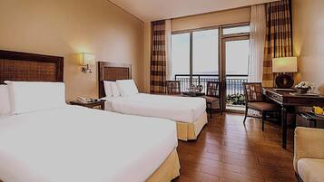A hotel room with two beds, a desk, a chair, a balcony with a view, and a window with curtains.