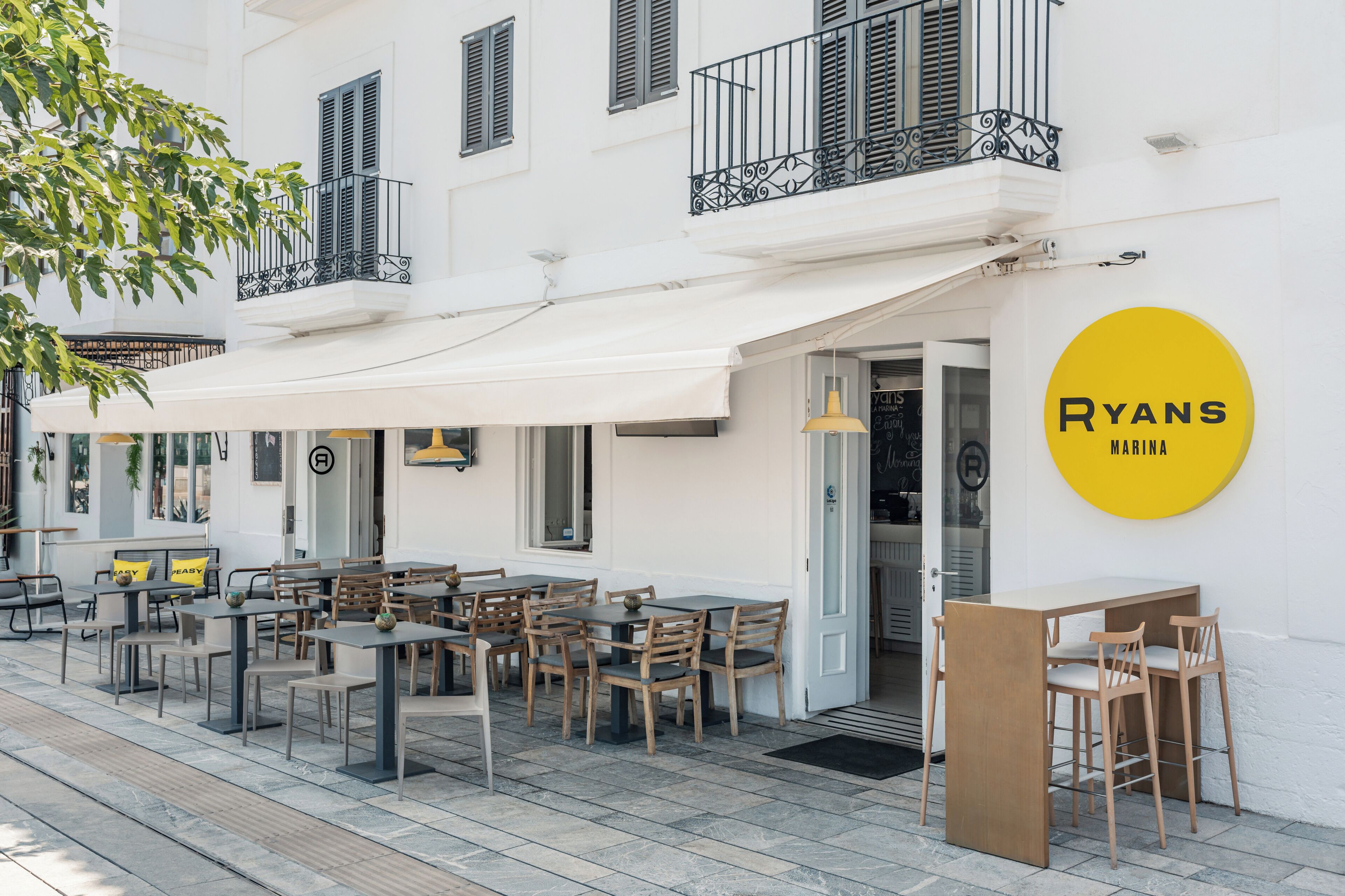An outdoor seating area with tables and chairs, a white awning, and a yellow sign for 'Ryans Marina'.