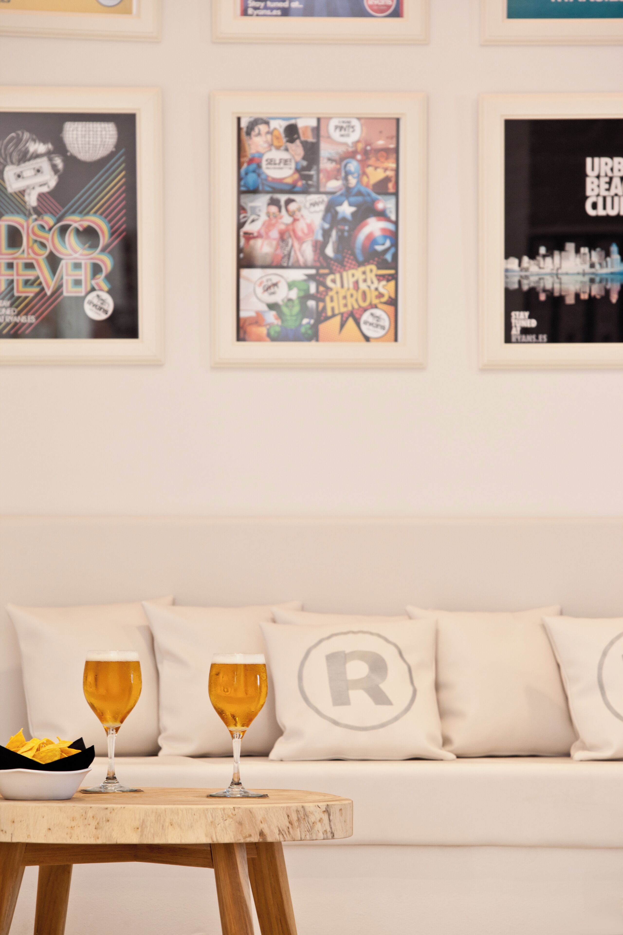 Een kamer met een witte bank, twee bierglazen op een houten tafel en lijstjes met posters aan de muur.