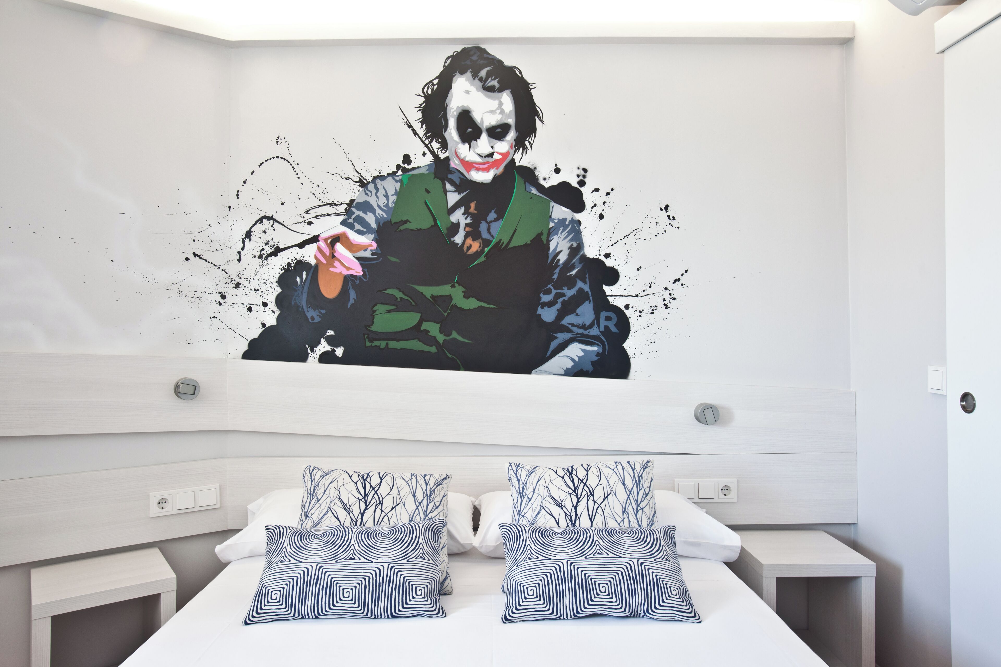 Ein modernes Schlafzimmer mit einer großen Wandmalerei einer Figur, einem ordentlich bezogenen Bett mit gemusterten Kissen und einem klaren, minimalistischen Design.