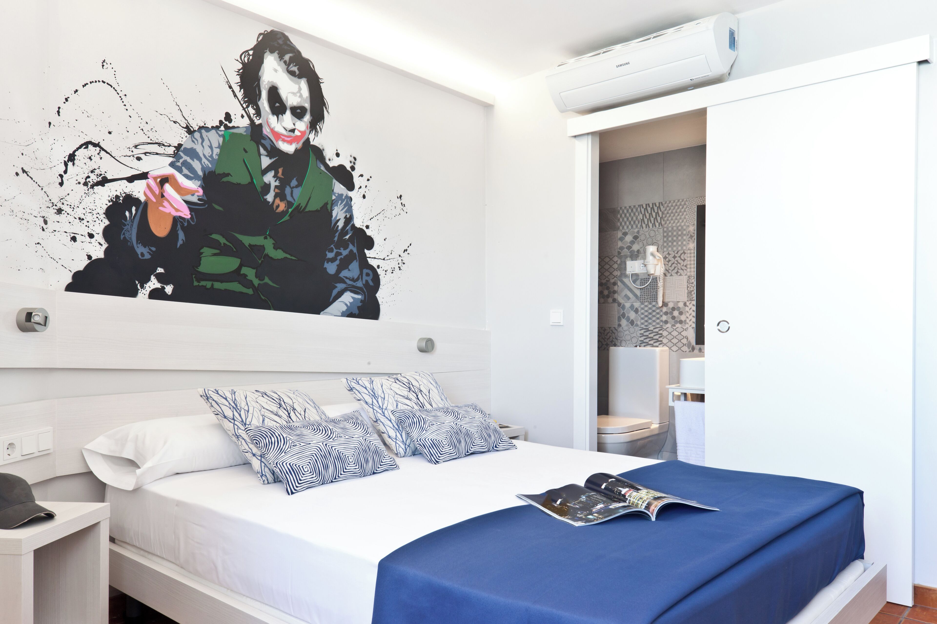 A modern hotel room with a large bed, a nightstand, a wall-mounted air conditioner, and a bathroom visible through an open door.