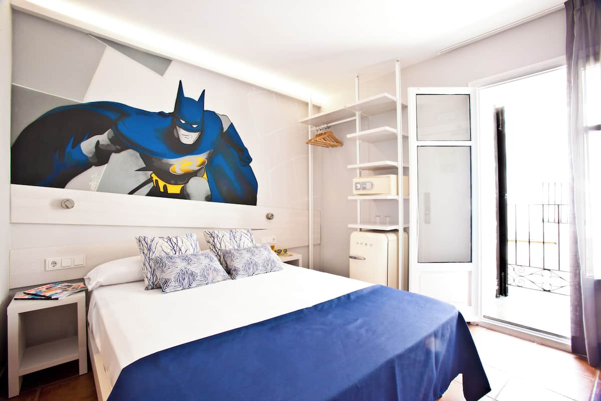 Ein Hotelzimmer mit einem großen Batman-Poster, einem ordentlich bezogenen Bett mit blauer Bettwäsche, einem kleinen Nachttisch mit einer Zeitschrift und einem Kleiderschrank mit Spiegel.