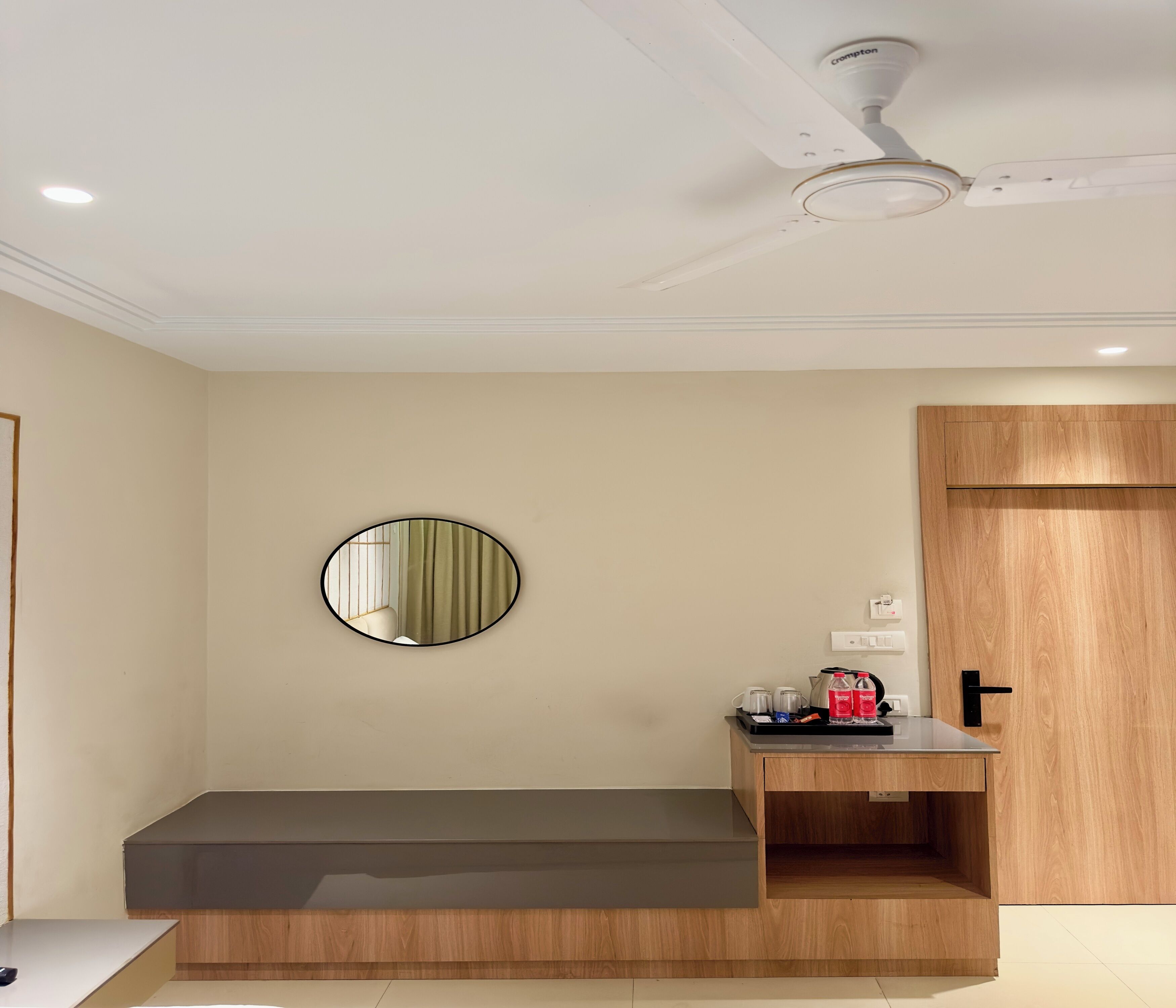 Una habitación moderna con un banco de madera, un espejo redondo, un ventilador de techo y una pequeña mesa de madera con una cafetera y botellas.