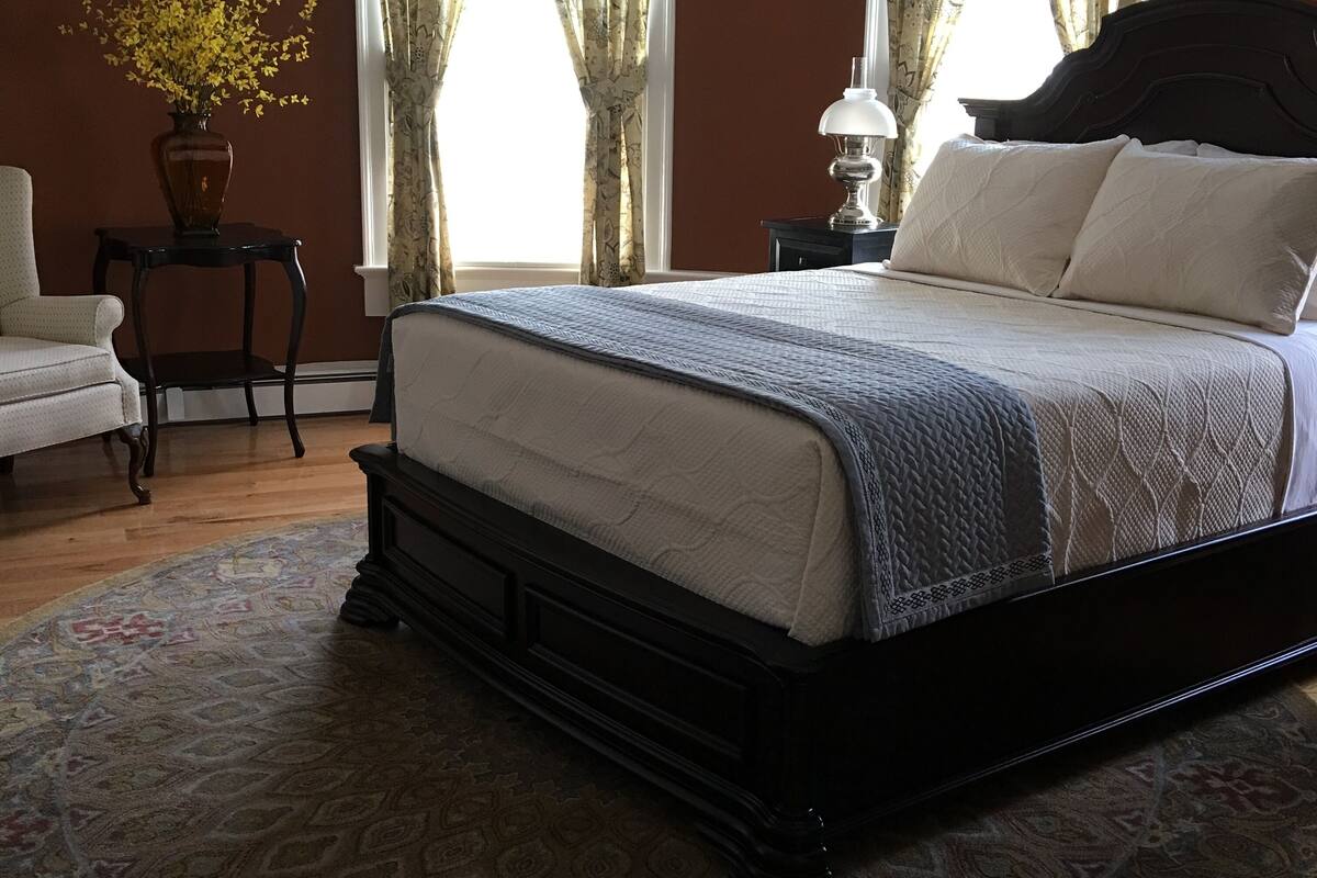 A bedroom with a large bed, a nightstand, a lamp, a chair, and a vase with flowers.