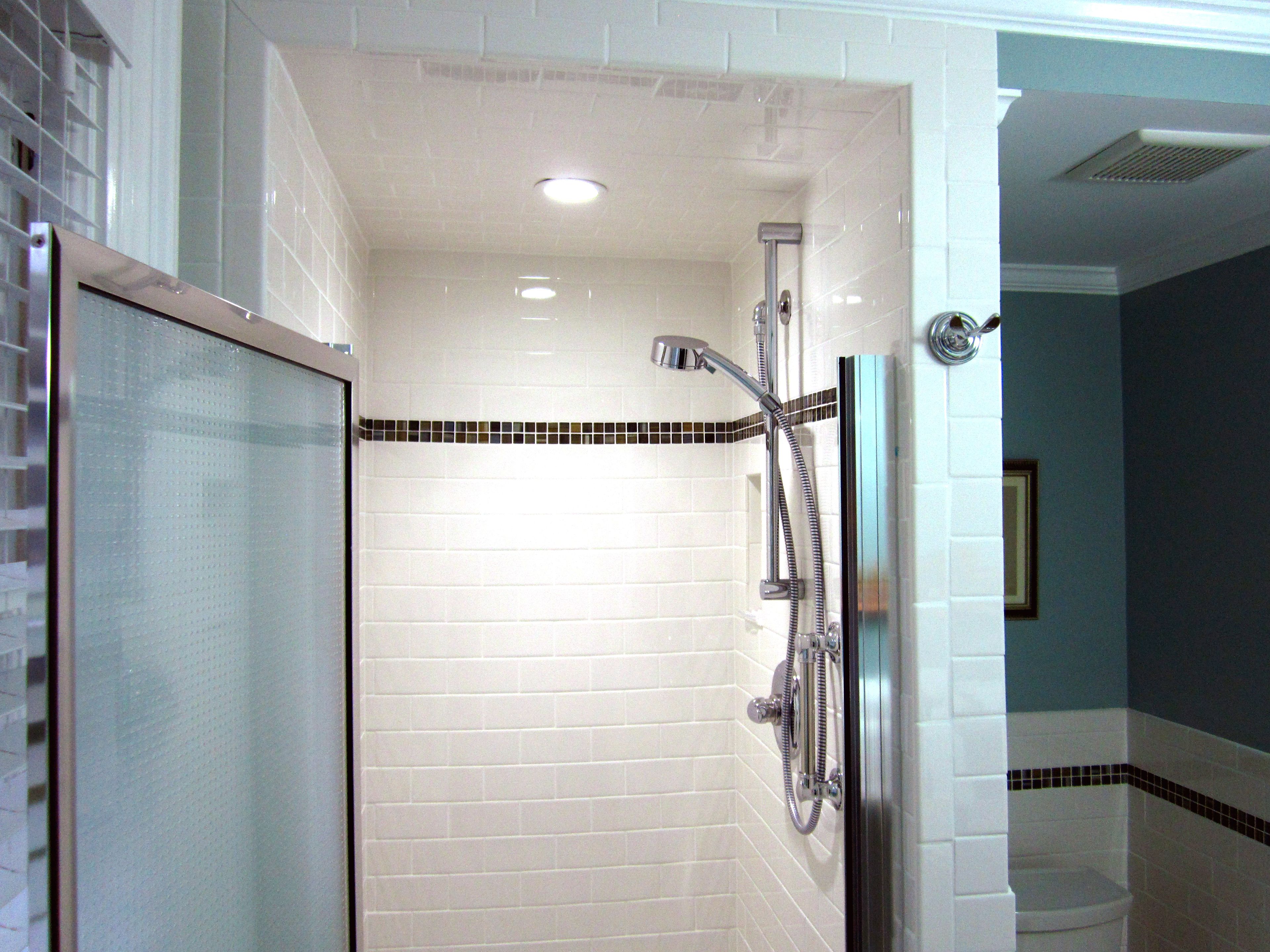 A bathroom with white tiled walls, a shower with a handheld showerhead, and a glass door.