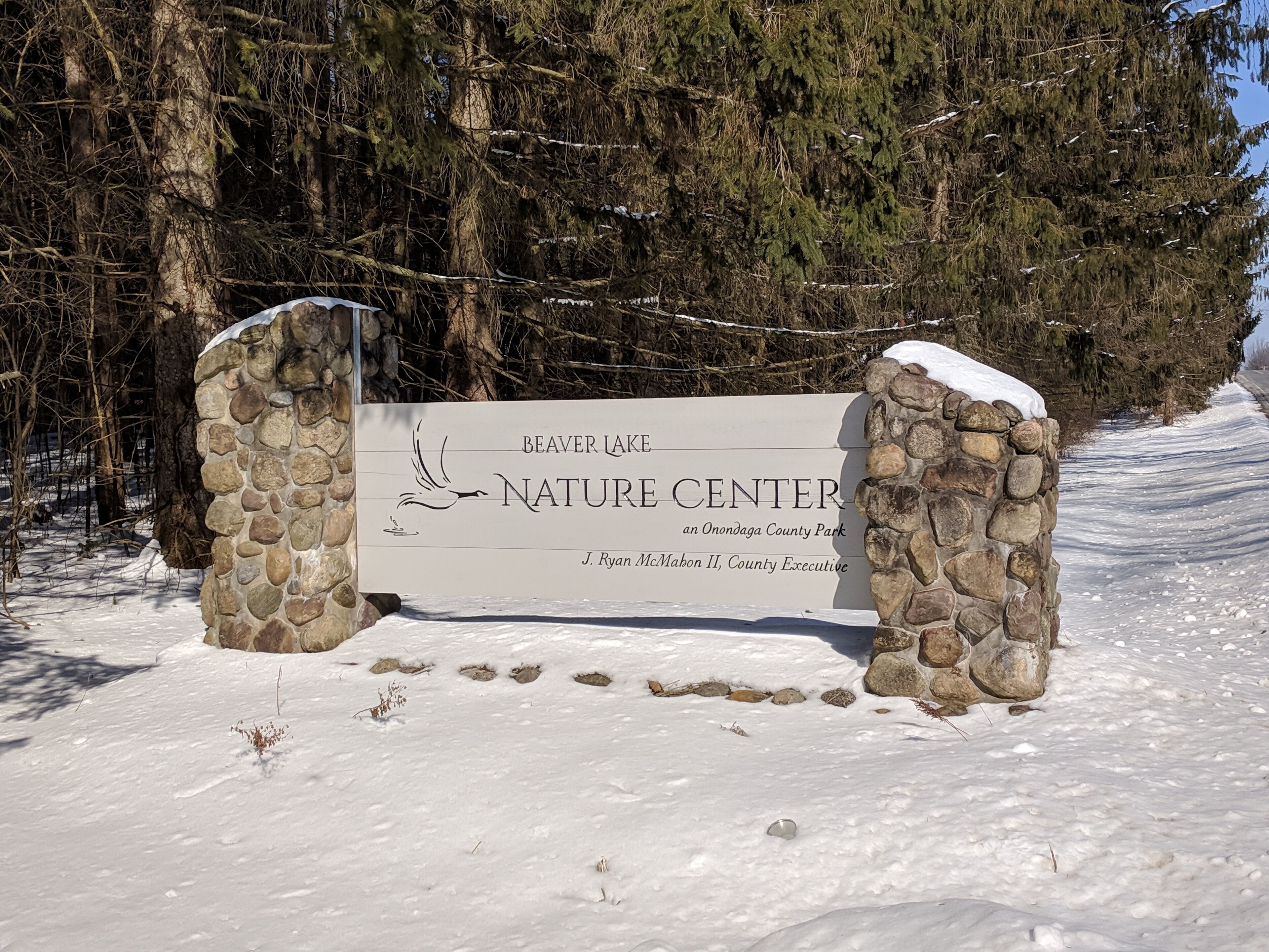 A stone sign for Beaver Lake Nature Center with a bird illustration, surrounded by snow and trees.