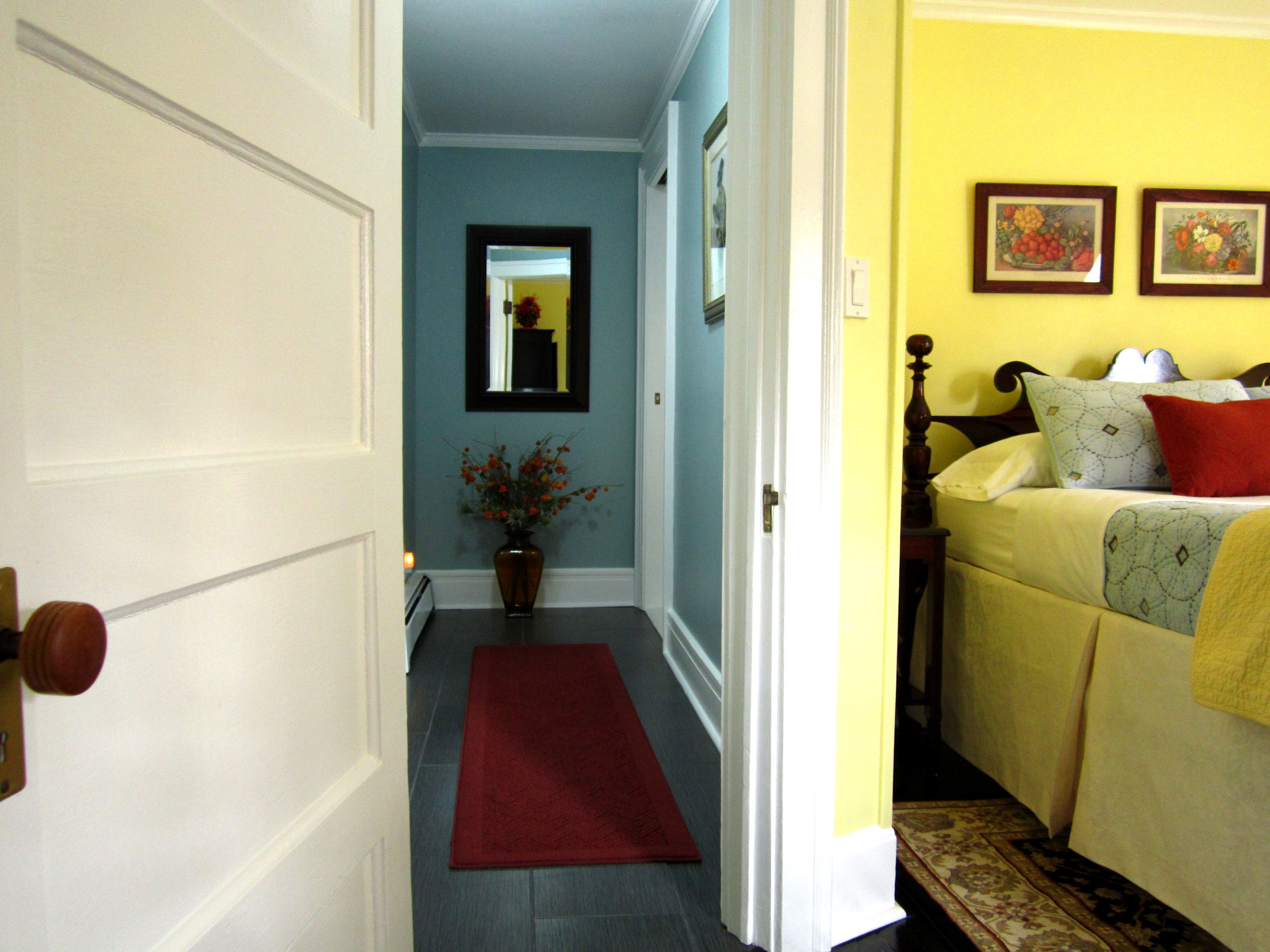 A hallway with a red rug leading to a bedroom with a bed, a nightstand, and a wall with two framed pictures.