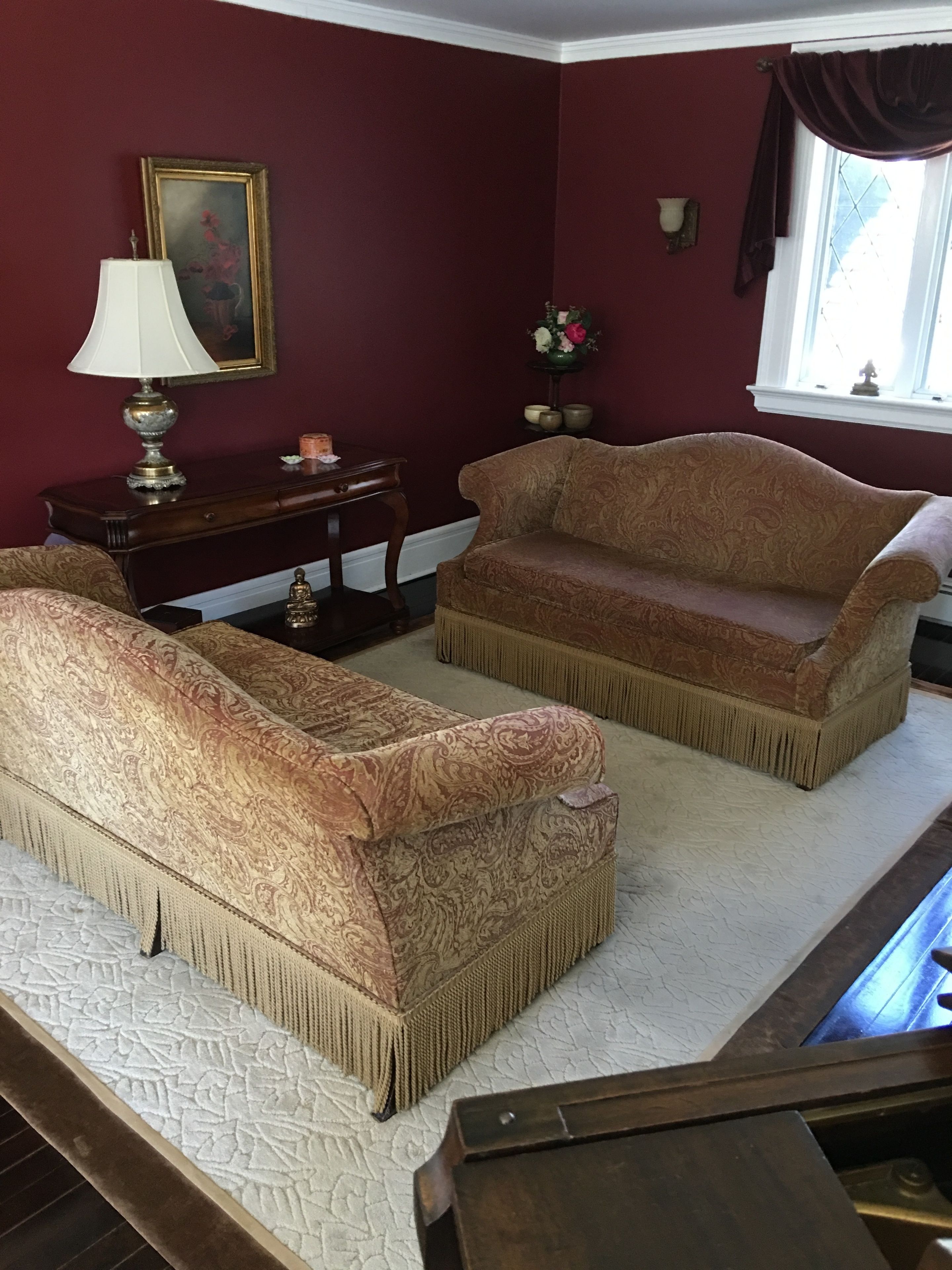 A room with two patterned sofas, a small table with a lamp, a painting on the wall, and a window with curtains.