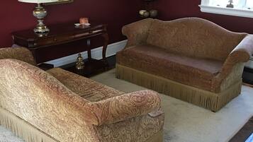 A room with two patterned sofas, a small table with a lamp, a painting on the wall, and a window with curtains.
