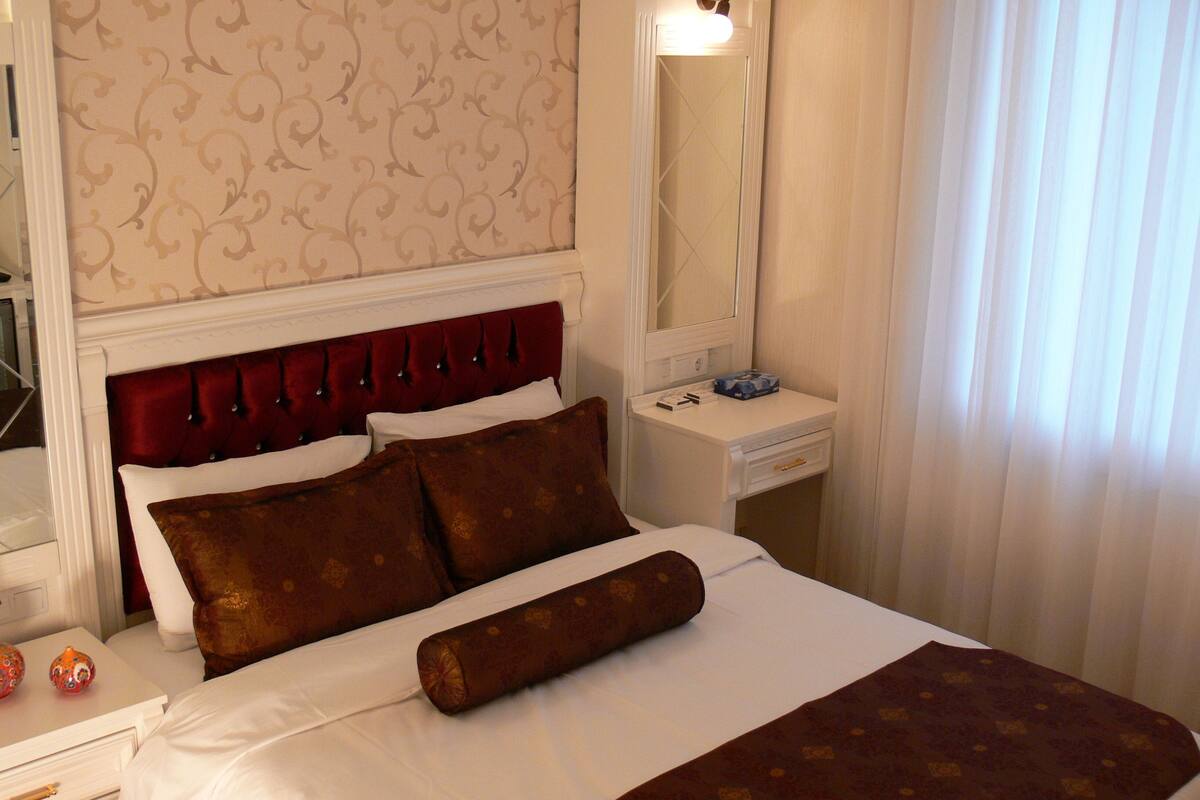 Una habitación de hotel con una cama grande, una mesita de noche, un aparador, un espejo y una ventana con cortinas.