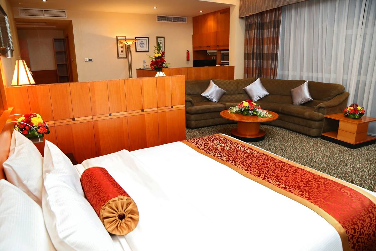Camera d'albergo con un letto, un divano, un tavolino con fiori e un angolo cottura sullo sfondo.