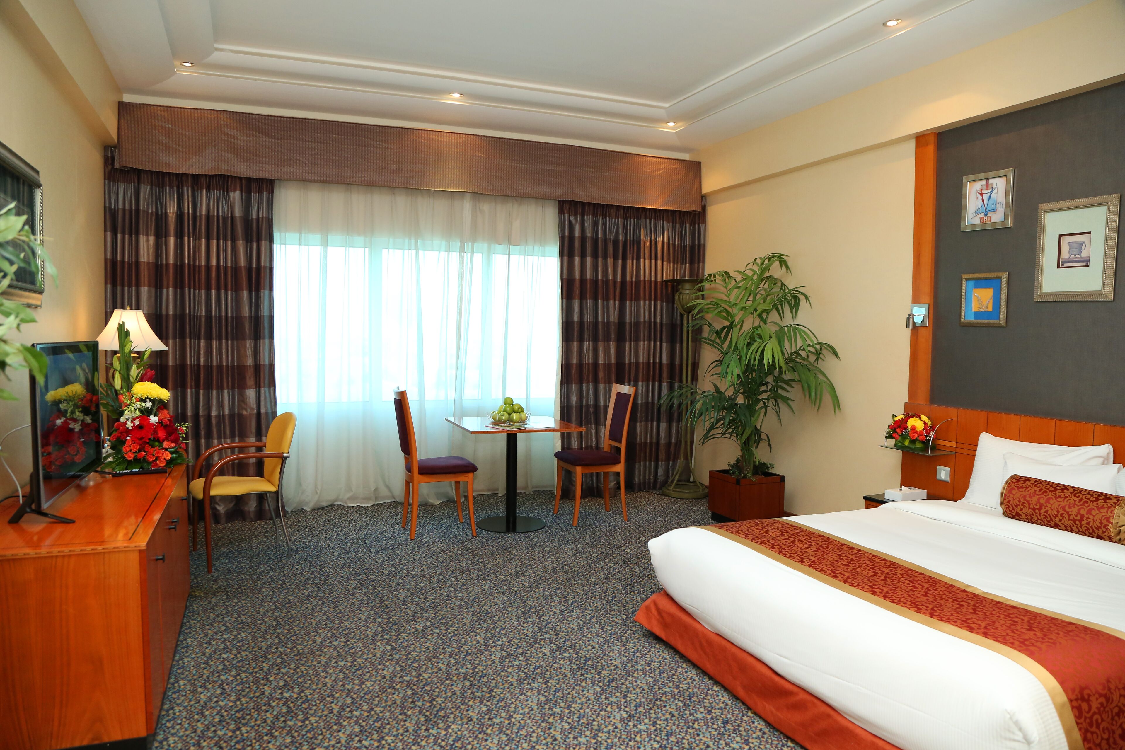 Une chambre d’hôtel avec un grand lit, deux chaises, une petite table ornée d’un vase de fleurs, un bureau avec une lampe et une grande plante en pot.