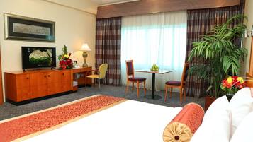 Habitación de hotel con cama, escritorio, silla, televisión, ventana con cortinas y una planta.