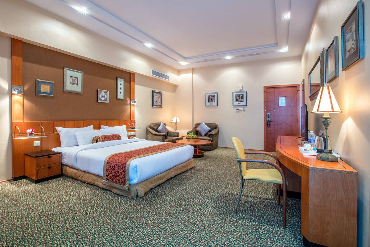 Una camera d'albergo con un letto grande, una scrivania, una sedia e un divano. Alle pareti si notano quadri incorniciati e sullo sfondo si vede una porta.