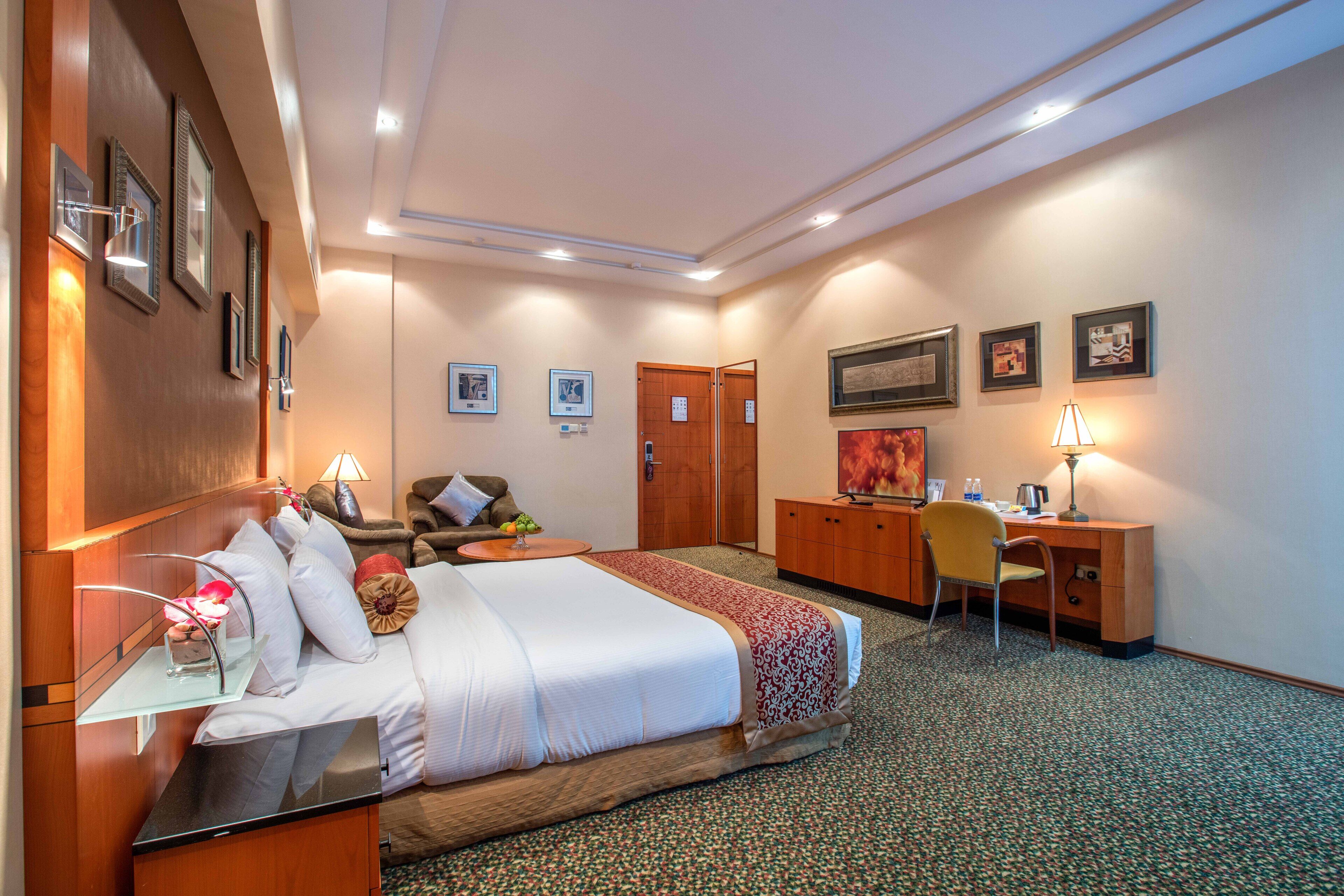 A hotel room with a bed, desk, chair, and a small table with a lamp.