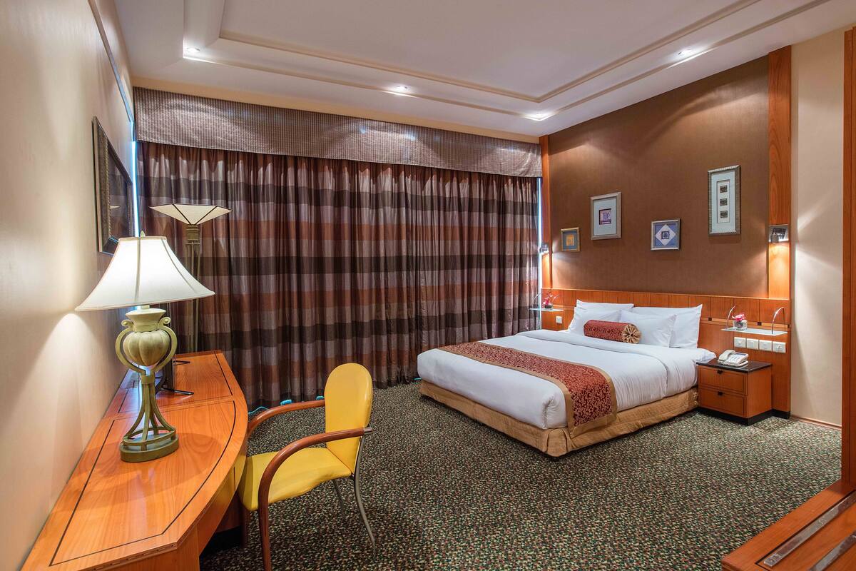 Una camera d'albergo con un letto, una scrivania con lampada, una sedia, un comodino e una parete con quadri.