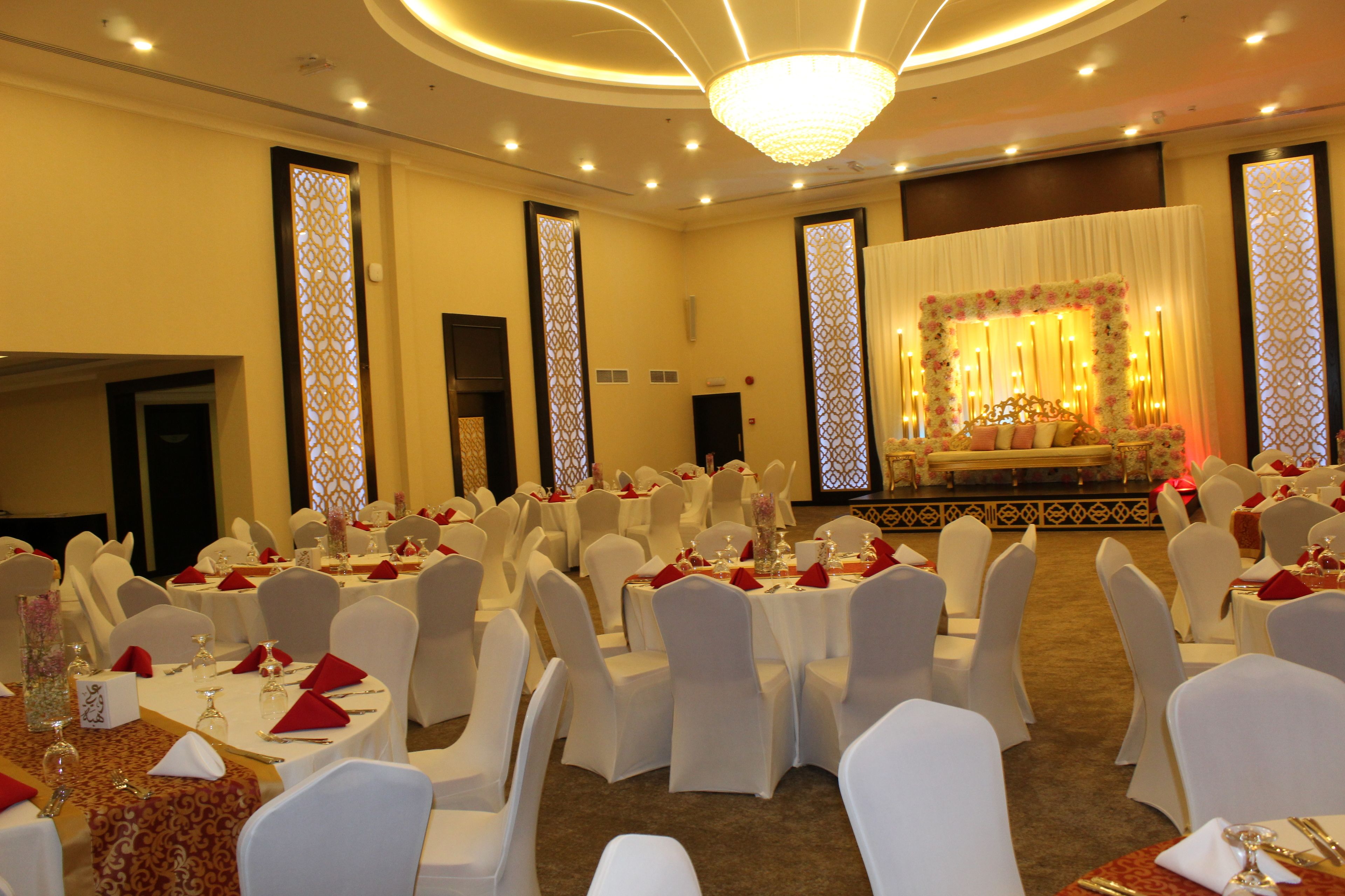 Un salón de banquetes con mesas redondas y sillas blancas, un escenario con decoración floral y una araña.