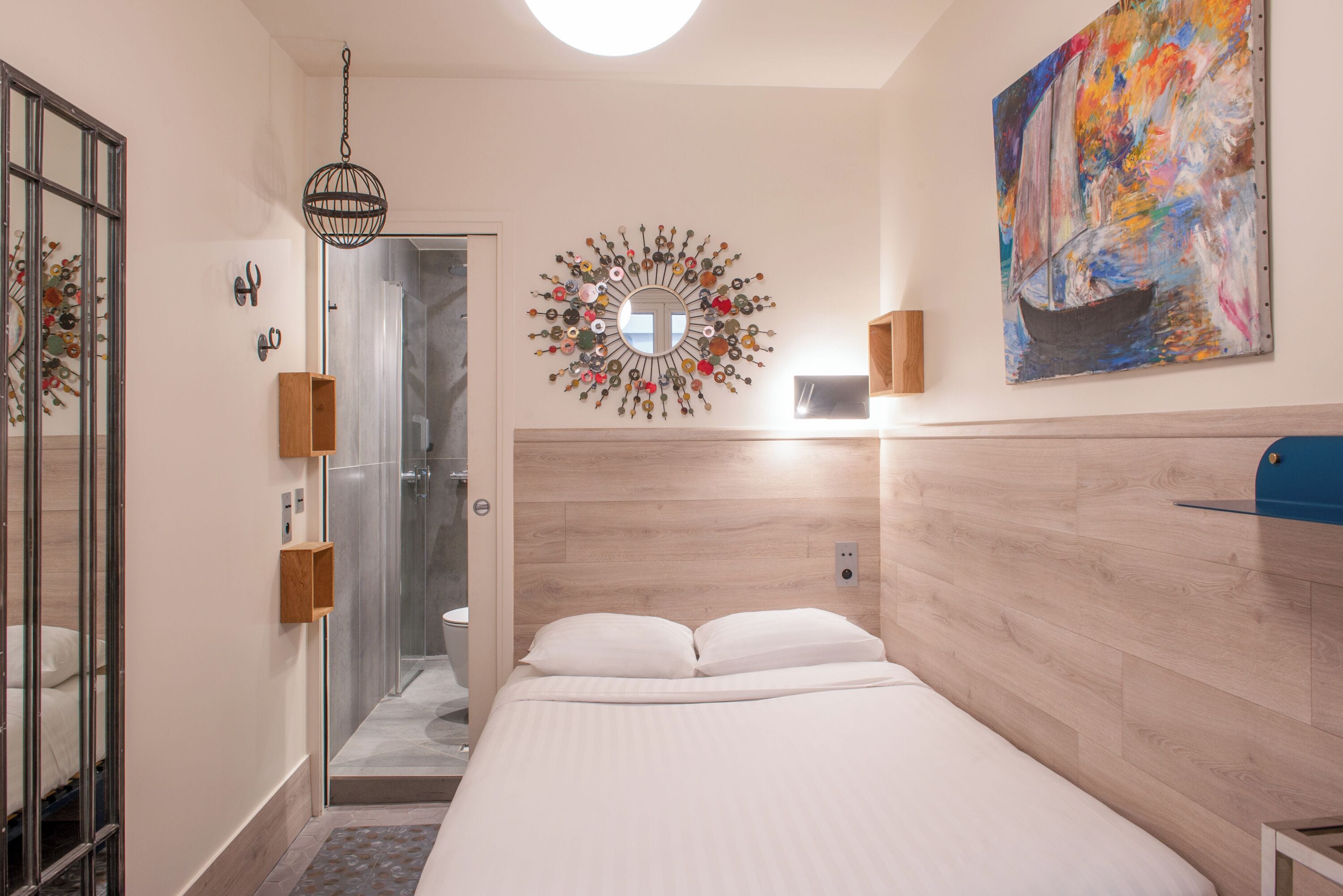 Una habitación de hotel moderna con cabecera de madera, una cama grande, zona de ducha y un cuadro abstracto colorido en la pared.