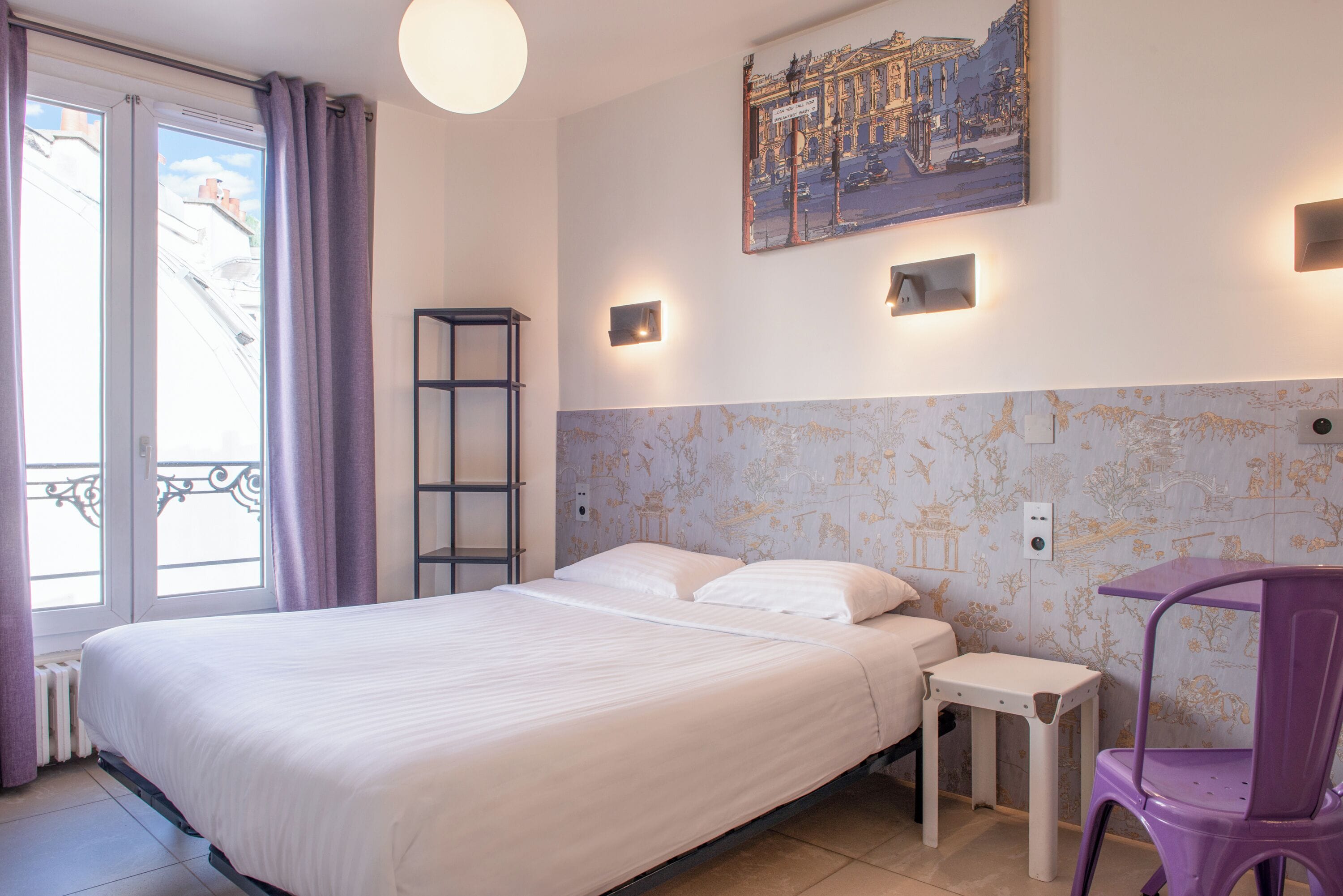 Une chambre avec un grand lit, une petite table, une chaise, un radiateur, une fenêtre avec des rideaux et un tableau accroché au mur.