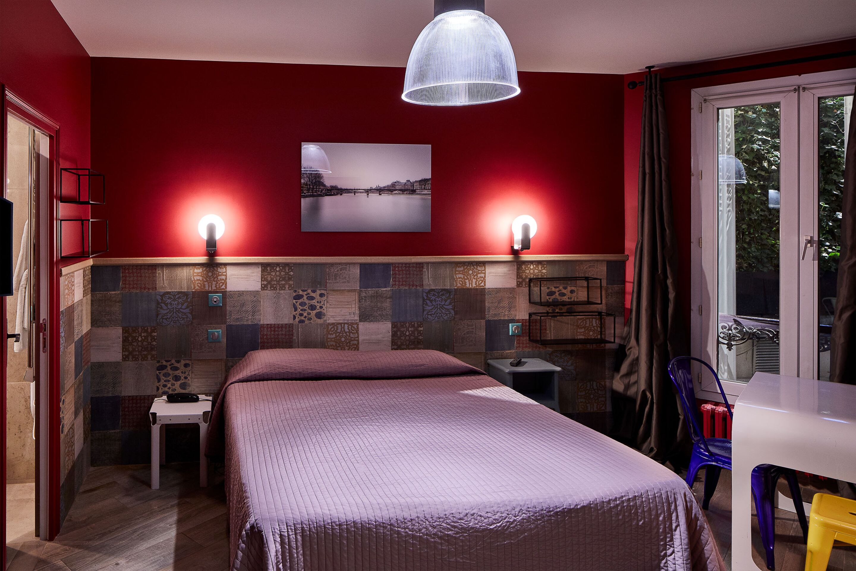 A hotel room with a bed, a table, a chair, a lamp, and a window with curtains.