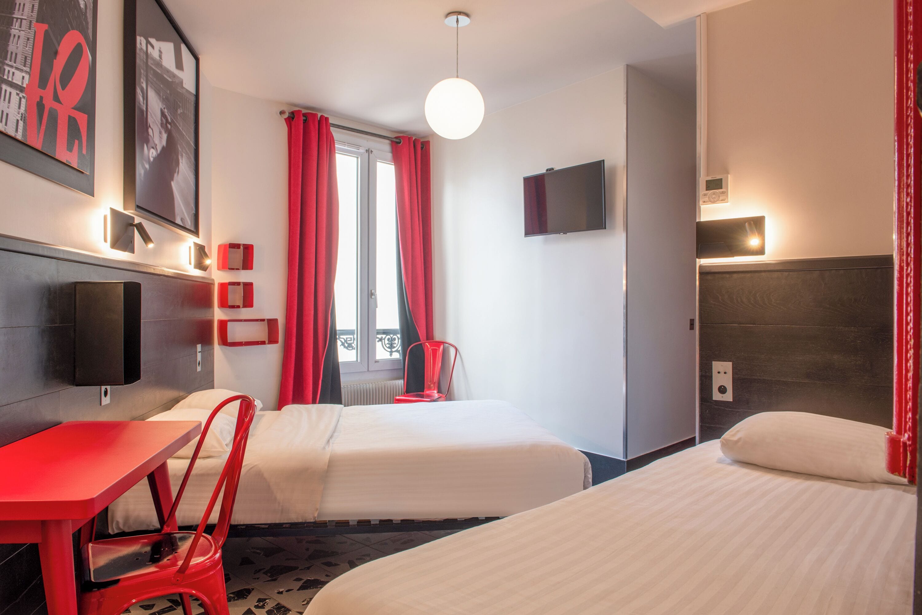 Ein Hotelzimmer mit zwei Betten, einem roten Tisch, einem roten Sessel, einem Fernseher und einem Fenster mit roten Vorhängen.