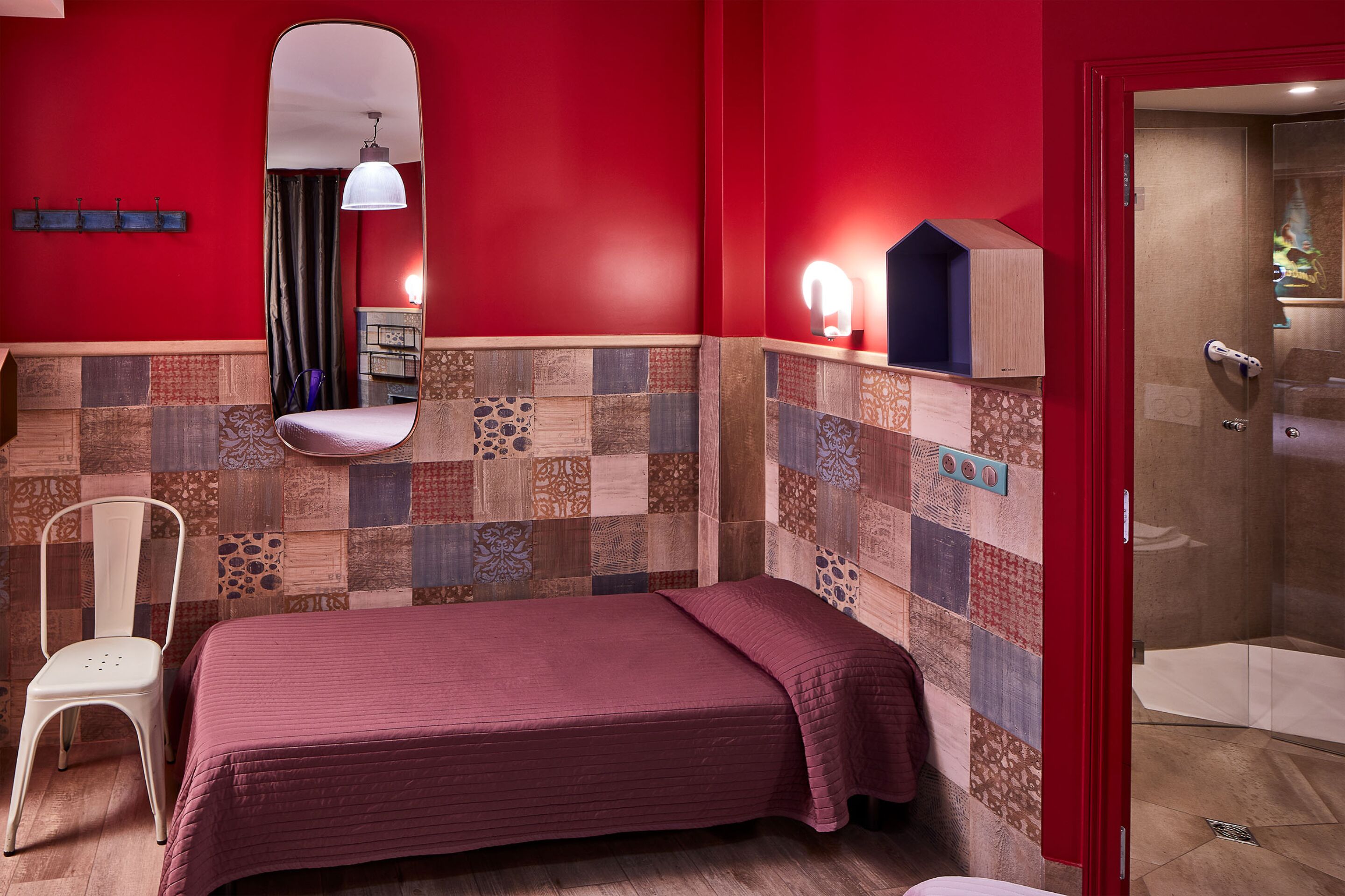 A hotel room with a red wall, a bed with a red bedspread, a chair, a mirror, and a shower area.