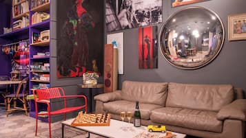 Una habitación con un sofá color beige, una silla roja, un tablero de ajedrez, dos copas de vino y una botella de vino sobre una mesa.