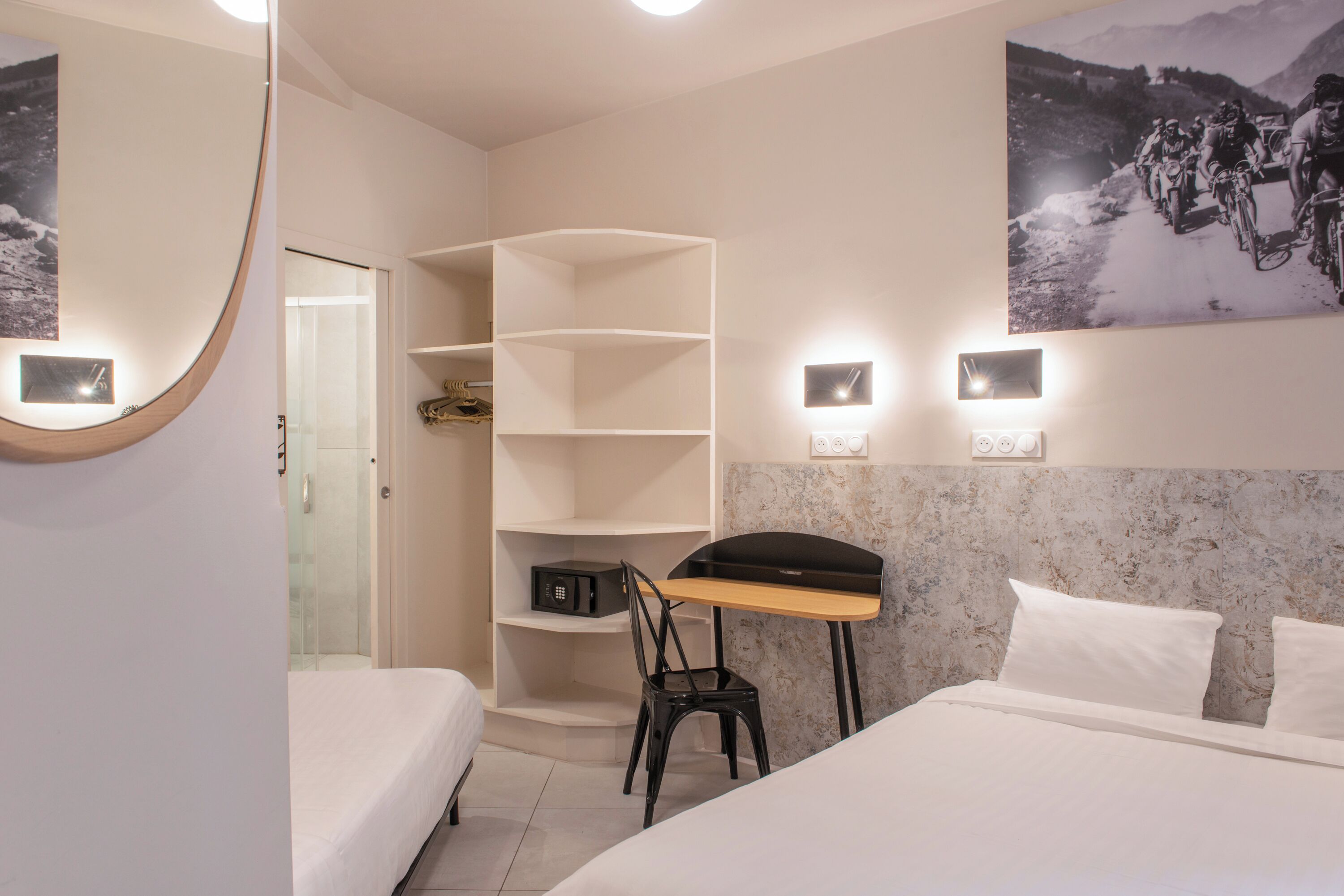Habitación de hotel con cama, escritorio, silla, estantería y lámpara de pared.