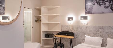 A hotel room with a bed, a desk, a chair, a shelf unit, and a wall-mounted light fixture.