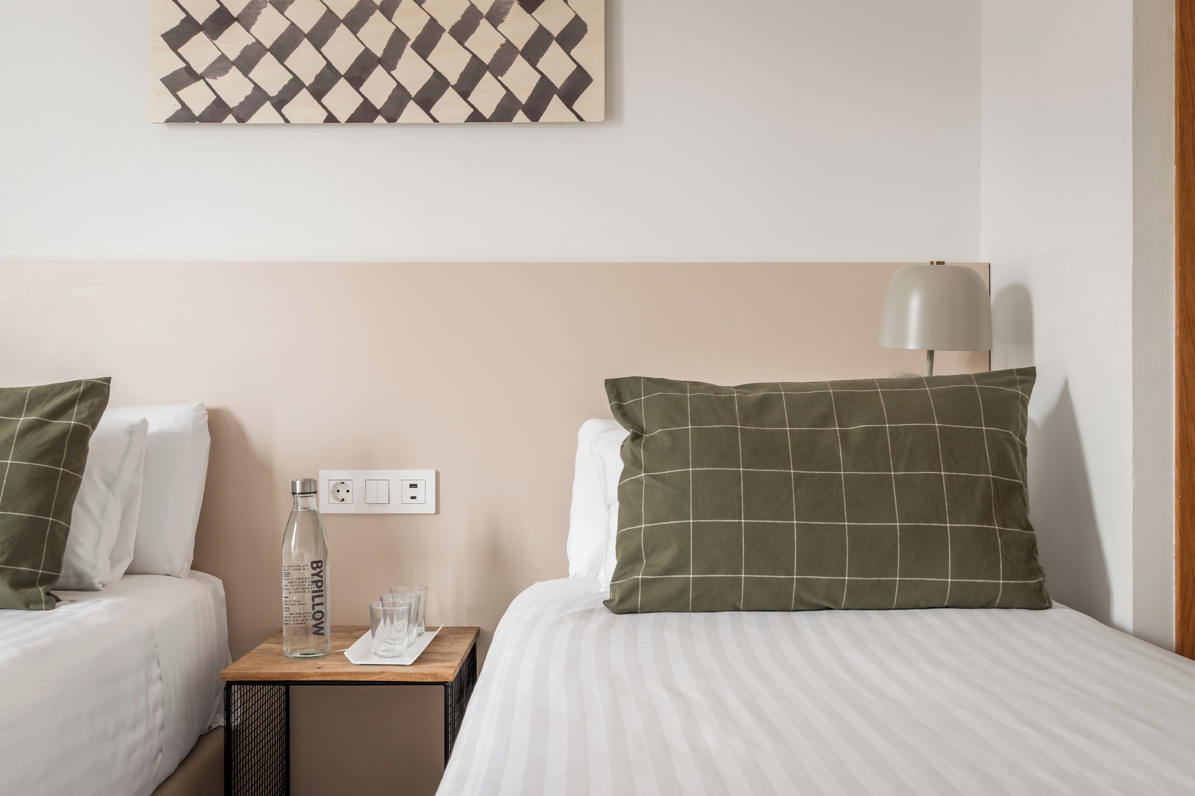Ein Hotelzimmer mit zwei Betten, einem Nachttisch mit Flasche und Gläsern, einer Lampe und einer Wandsteckdose.