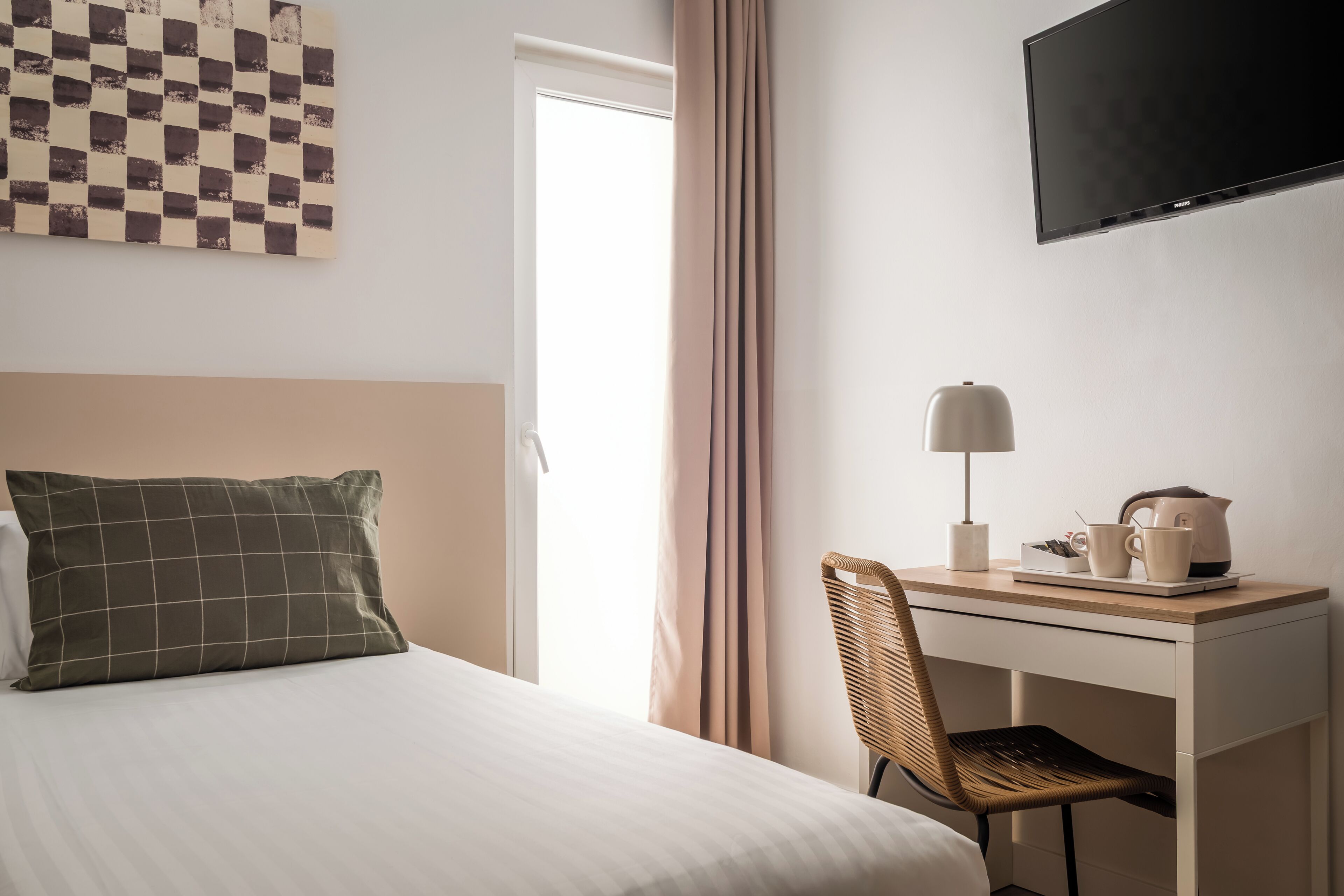 Habitación de hotel con una cama, un escritorio con lámpara, una silla, un televisor y un cuadro de pared a cuadros.