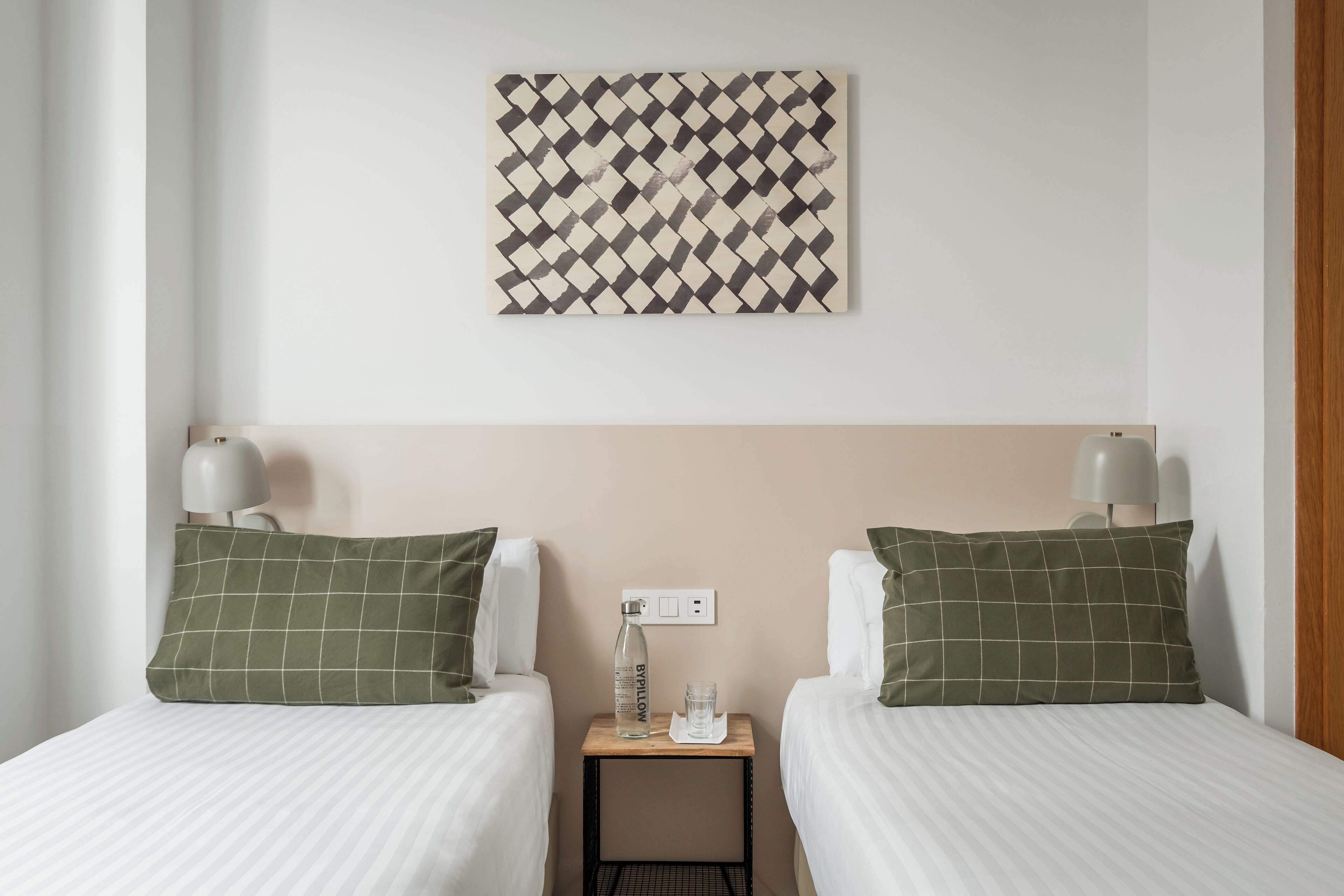 Une chambre d’hôtel avec deux lits, chacun agrémenté d’un oreiller à carreaux verts et d’un oreiller blanc, une petite table avec une bouteille et un verre, et un interrupteur fixé au mur.