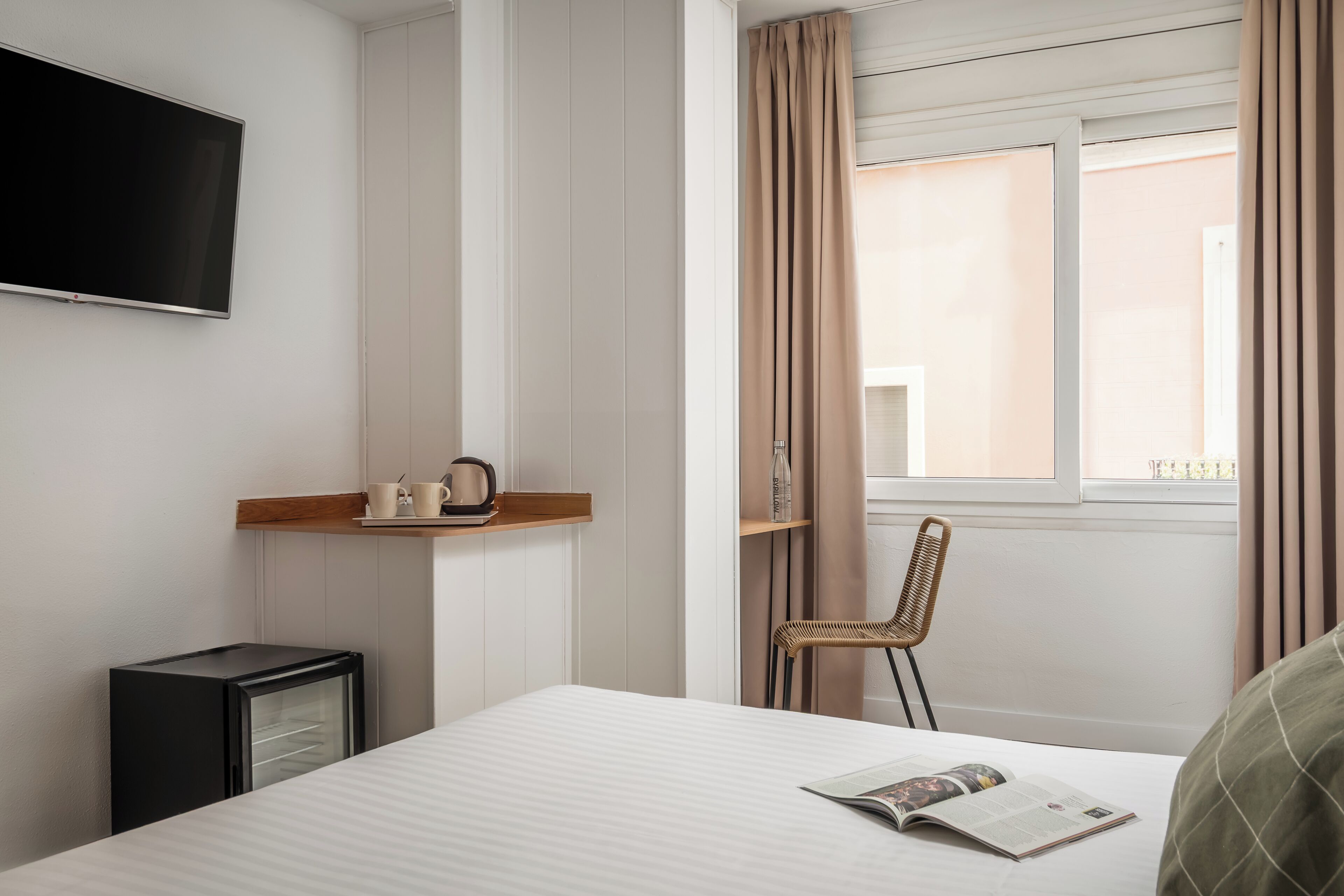 Habitación de hotel con cama, un refrigerador negro pequeño, un televisor de pantalla plana montado en la pared, un estante de madera con una tetera y tazas, una silla de mimbre y una ventana con cortinas.
