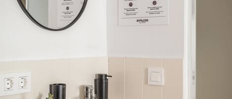 Un baño moderno con un lavabo blanco, un espejo redondo y un grifo empotrado en la pared. Hay un cartel con consejos para ahorrar agua, una planta pequeña en maceta y un interruptor de luz en la pared.