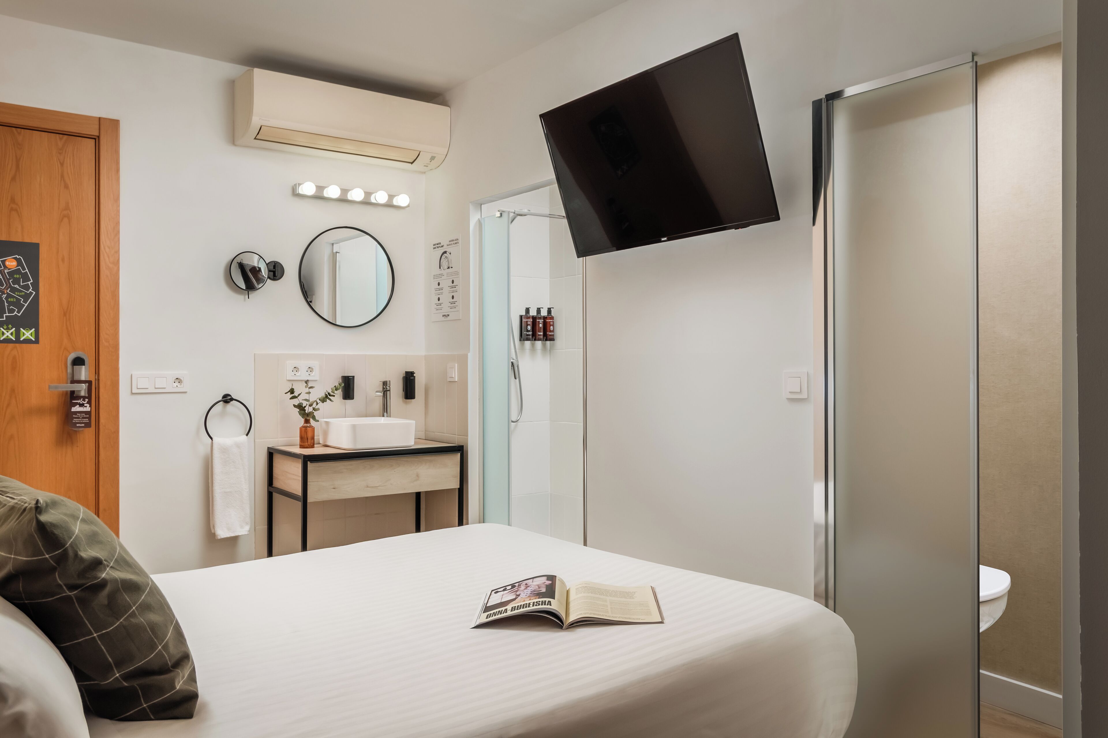 Una habitación de hotel con cama, mesita de noche con un libro, espejo, televisor montado en la pared y baño con inodoro y ducha.