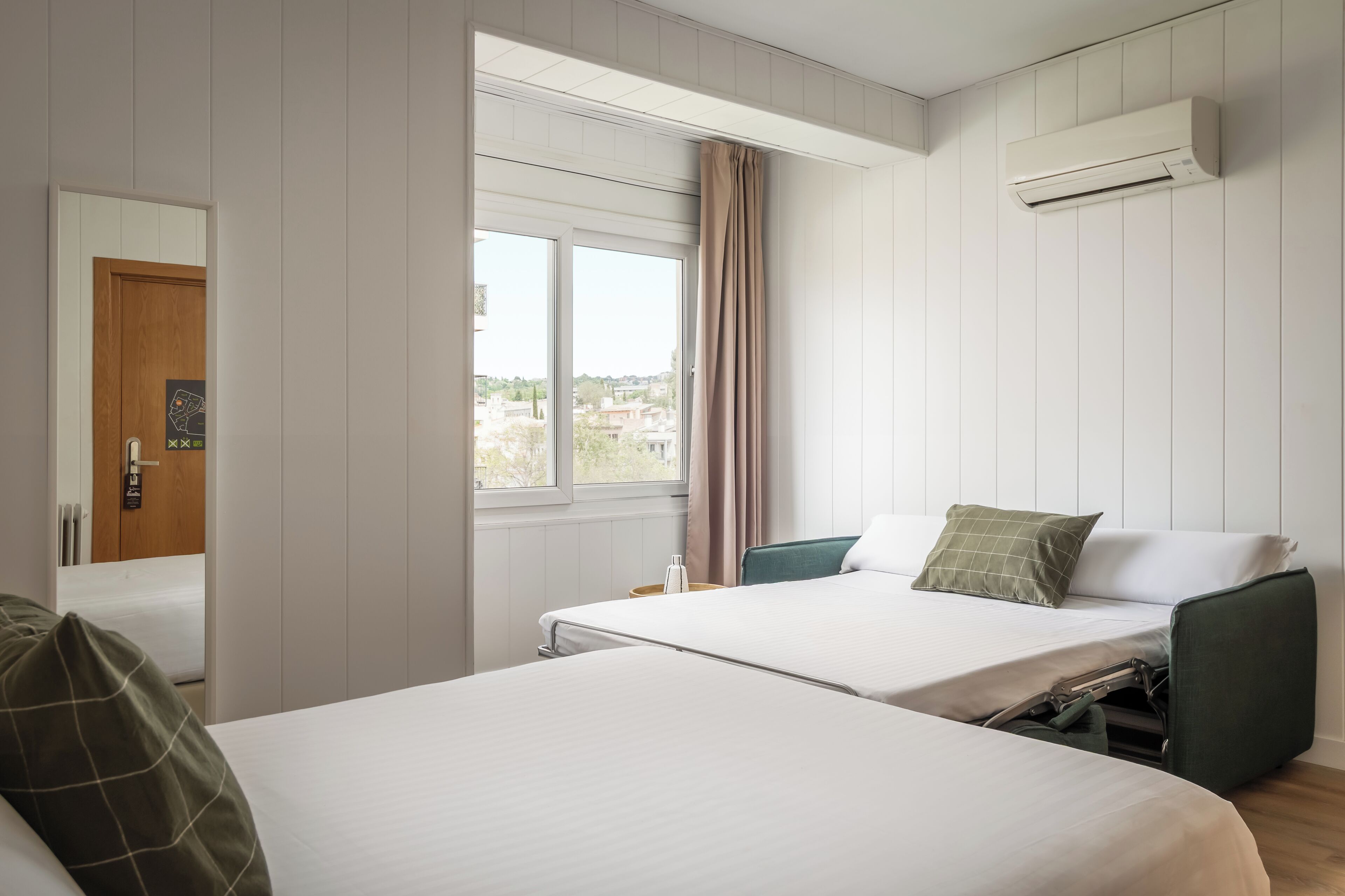 A hotel room with two beds, a window with curtains, an air conditioning unit, and a view of the city.