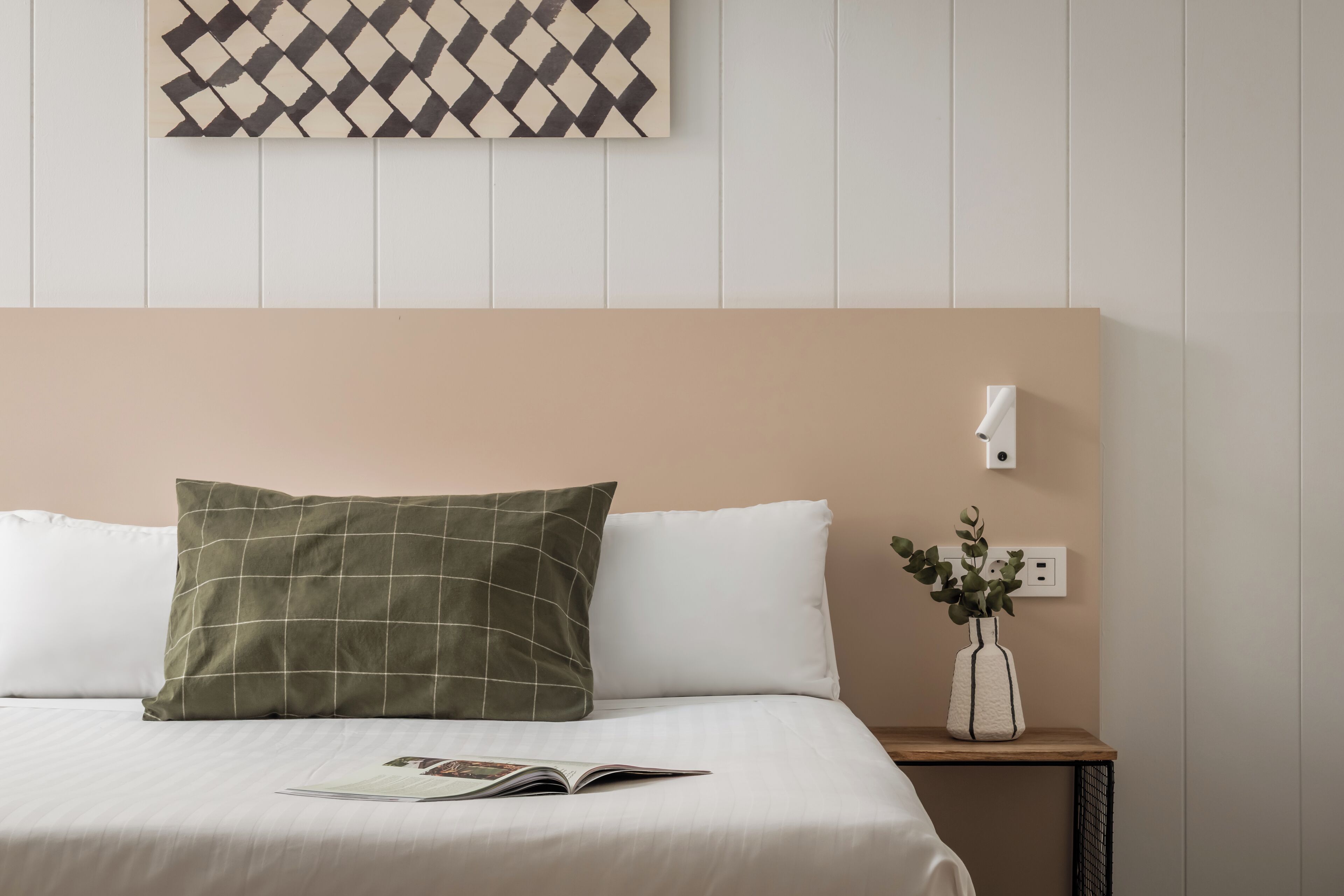A neatly made bed with a green and white pillow, a white pillow, and a magazine on the bed. There is a small nightstand with a white vase containing green plants.