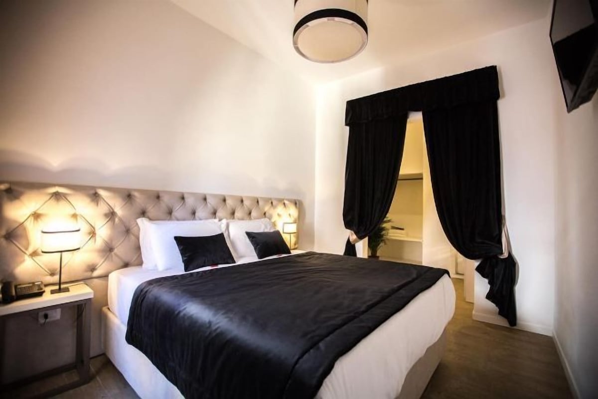 Un dormitorio con cabecera capitonada, una cama grande con ropa de cama blanco y negro, una mesita de noche con lámpara y cortinas negras en la ventana.
